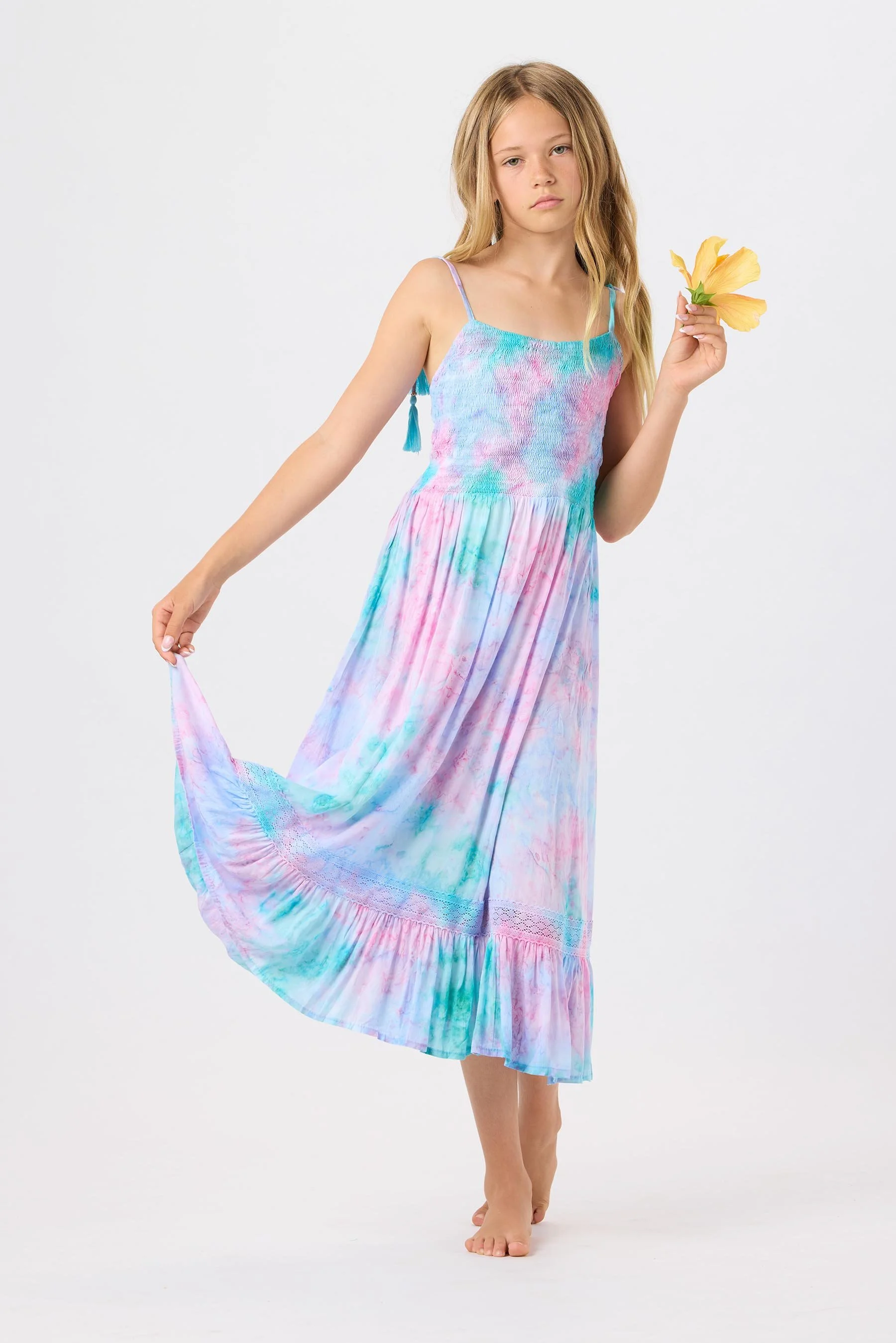 A young girl with long blonde hair wearing a colorful tie-dye maxi dress, holding a yellow flower, and standing against a white background.