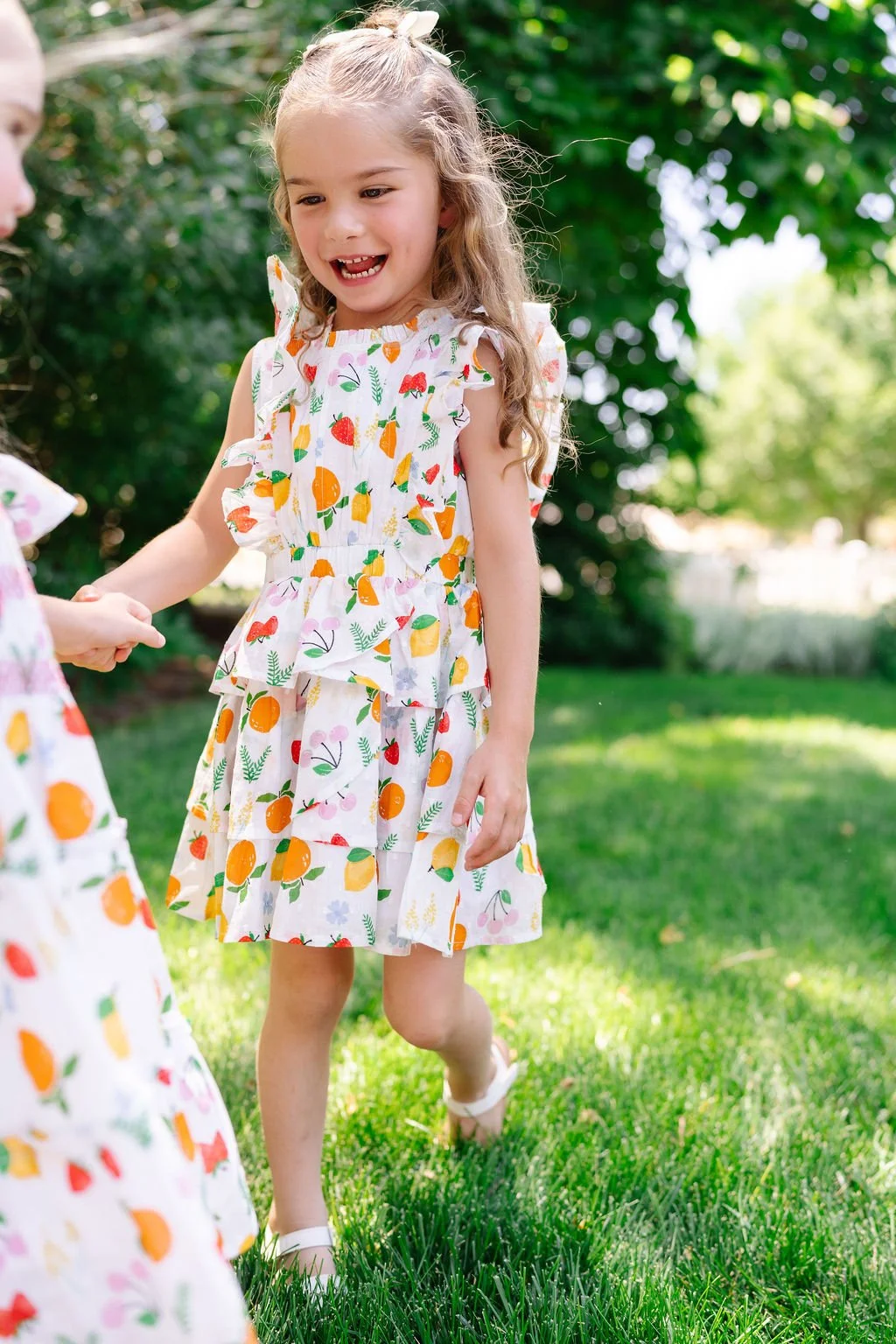 A young girl wearing a colorful, fruit-themed sundress holding hands with someone outside on a grassy area with trees in the background.