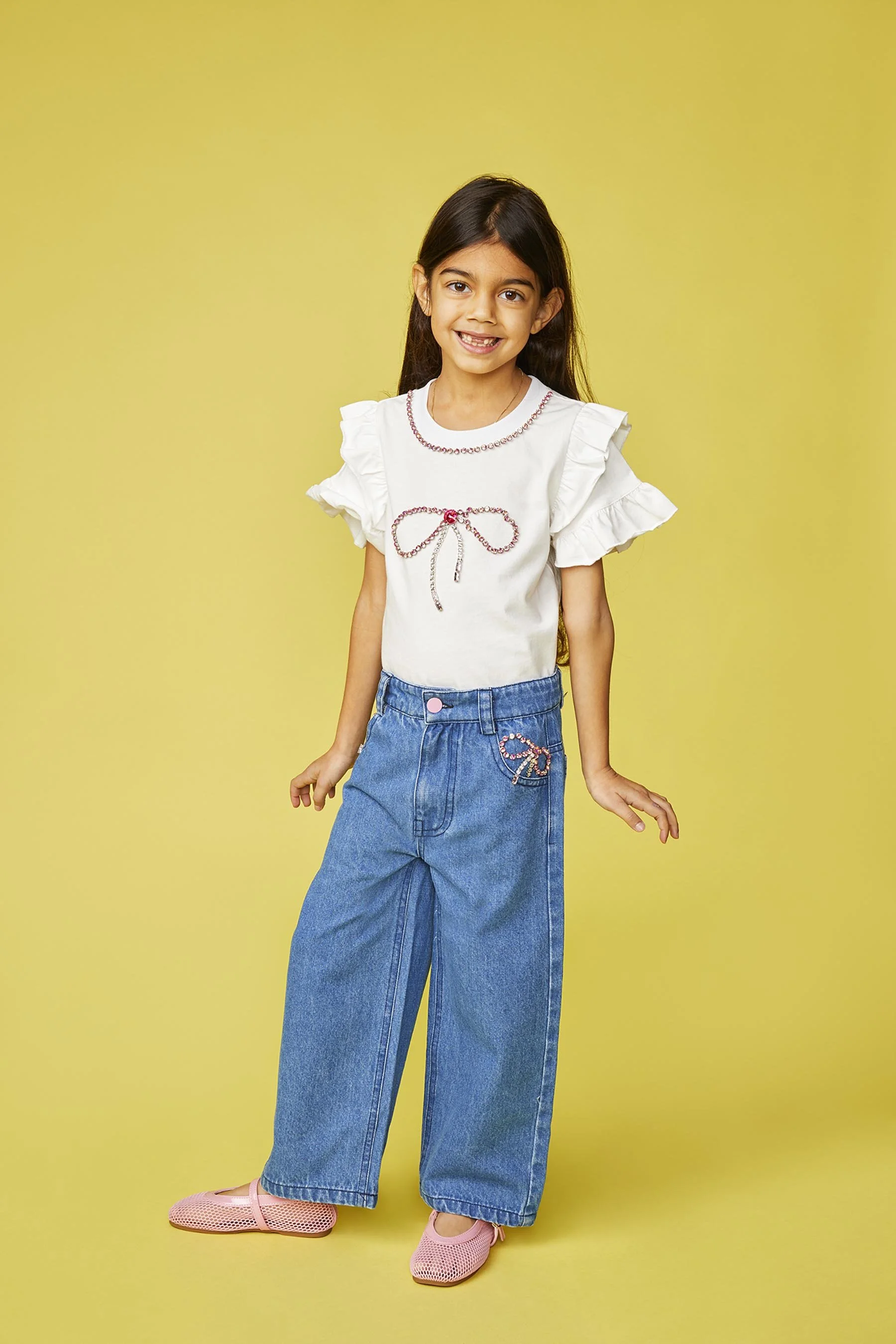 A young girl with long dark hair wearing a white ruffled sleeve top with a bow design, loose high-waisted blue jeans with an embroidered bow, and pink shoes, standing against a yellow background and smiling.