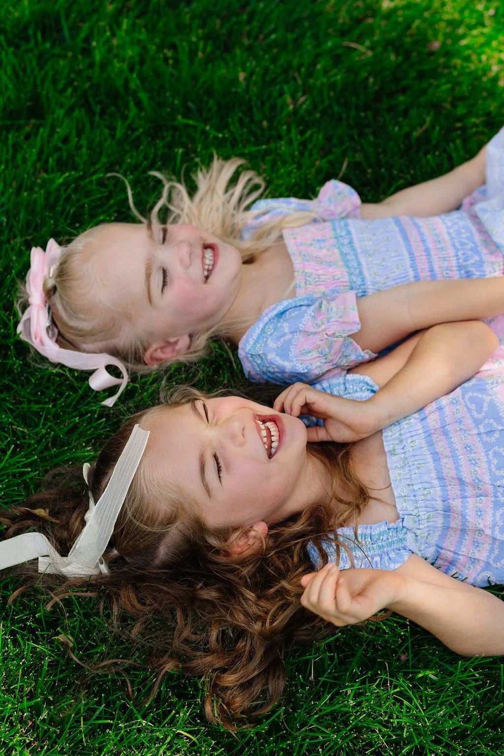 Two young girls with long hair and bows lying on green grass, laughing and smiling.