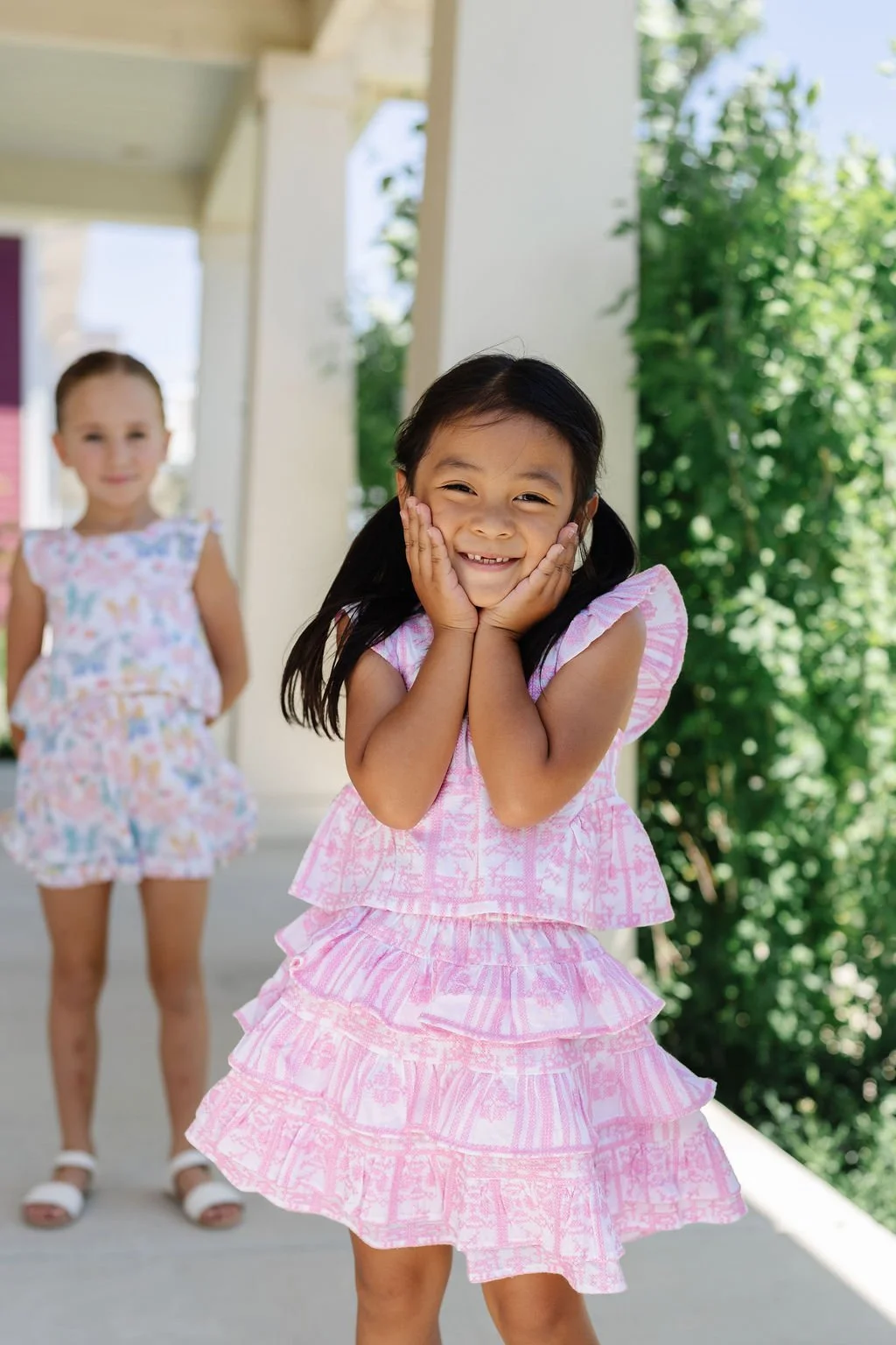 Two young girls in colorful summer dresses smiling outdoors on a porch with greenery in the background.