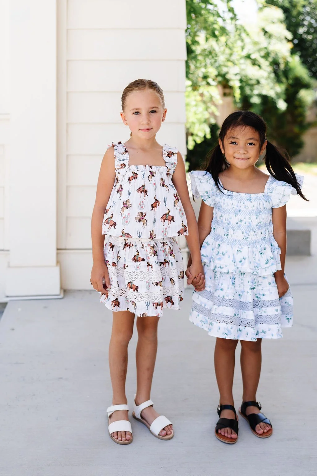 Two young girls holding hands outside, wearing summer dresses with ruffled sleeves and floral patterns, smiling in front of a white house and green trees.