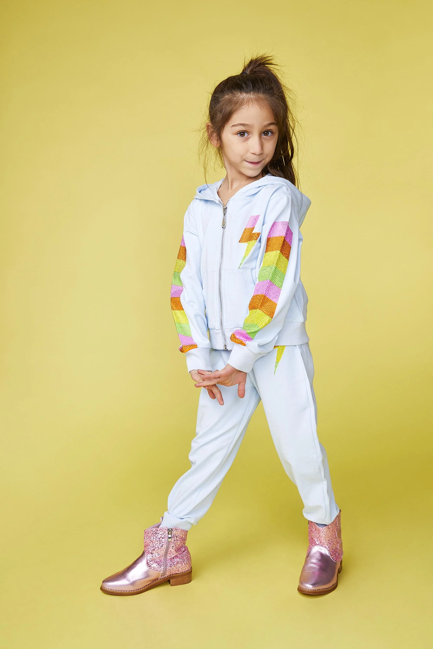A young girl with long brown hair in a high ponytail, standing against a yellow background, wearing a light blue tracksuit with rainbow-colored lightning bolt patterns on the sleeves and chest, and pink glittery boots.