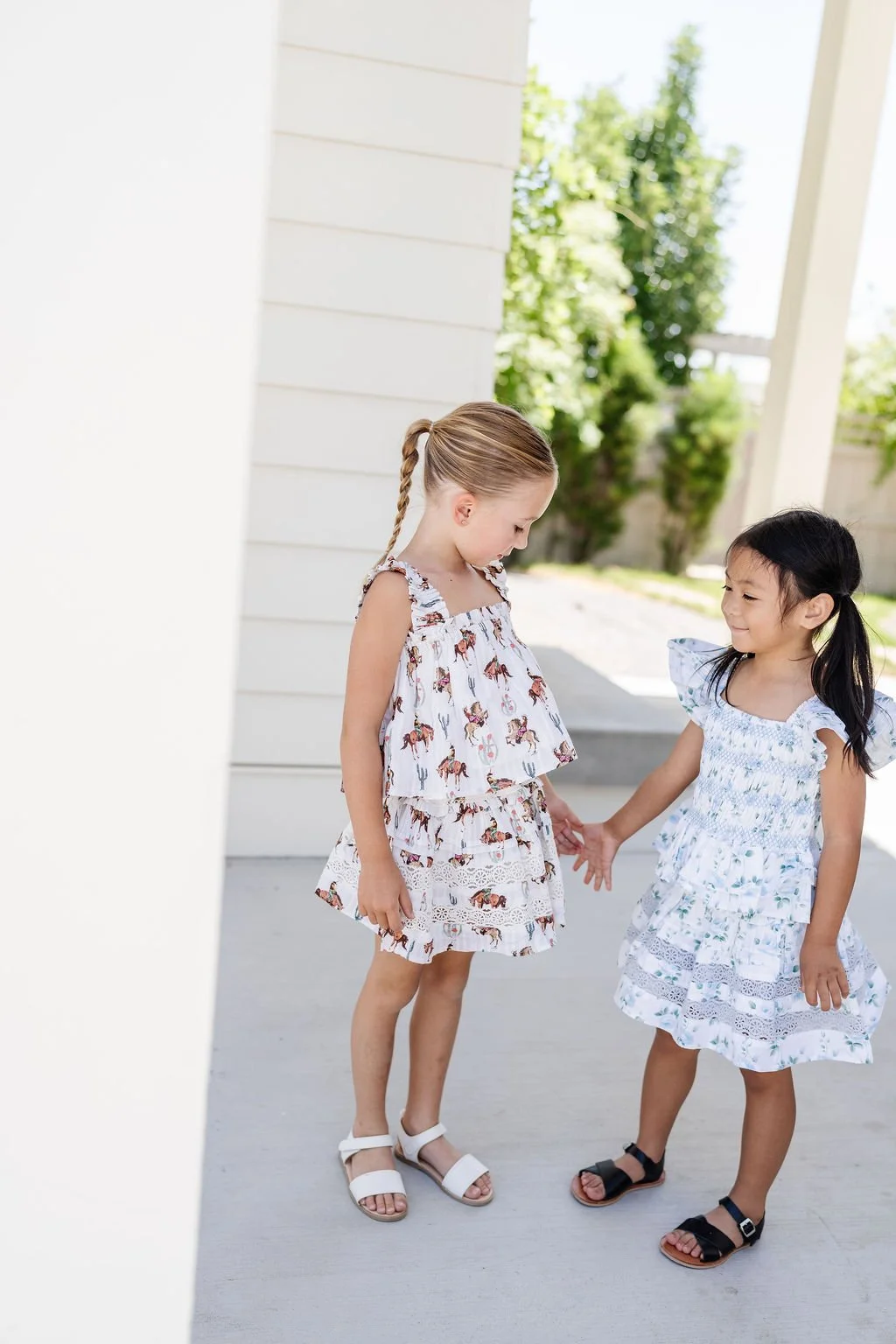 Two young girls holding hands and smiling outside on a porch, wearing summer dresses and sandals.
