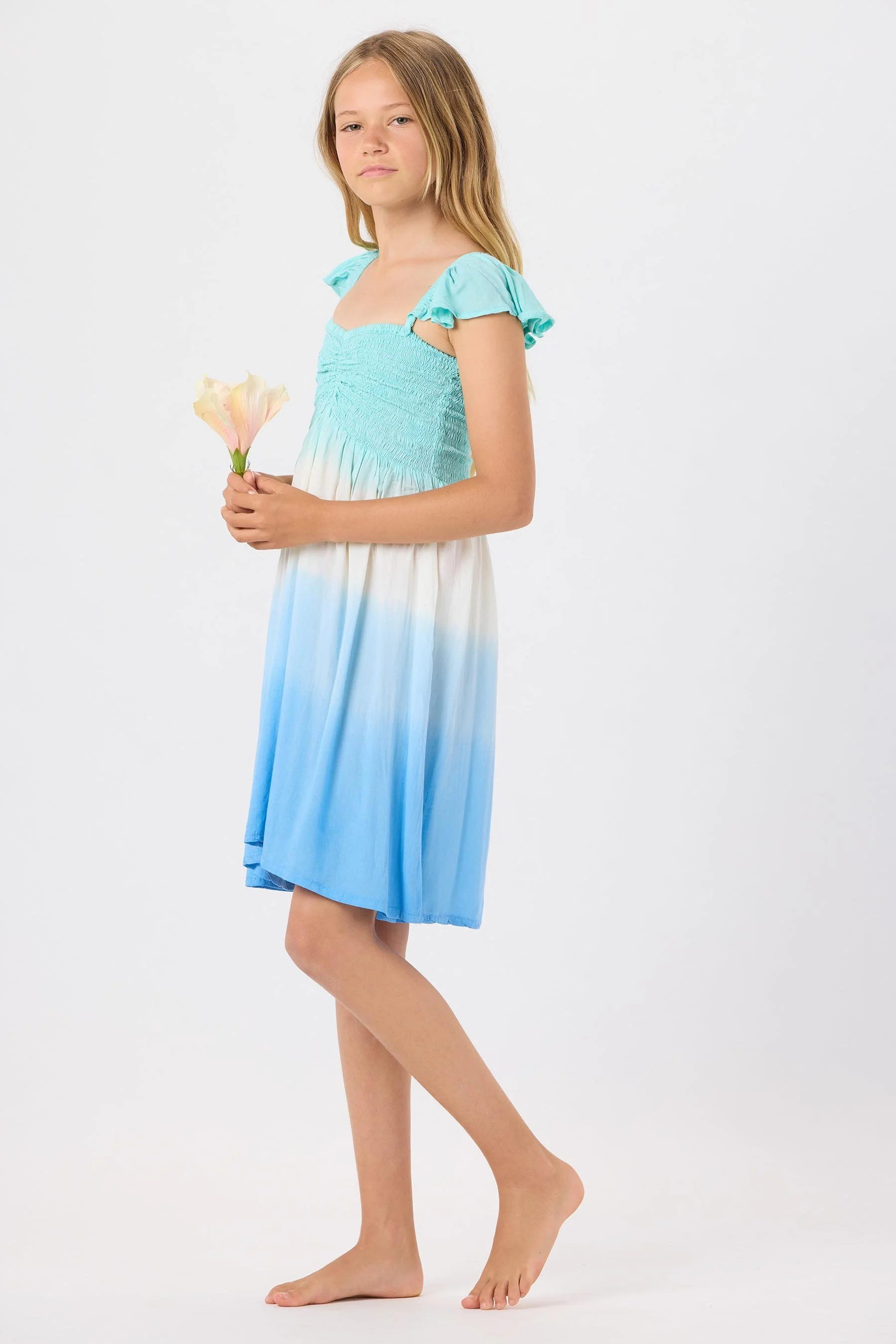 Young girl with light brown hair wearing a blue and white gradient dress, holding a pink flower, standing barefoot against a plain white background.