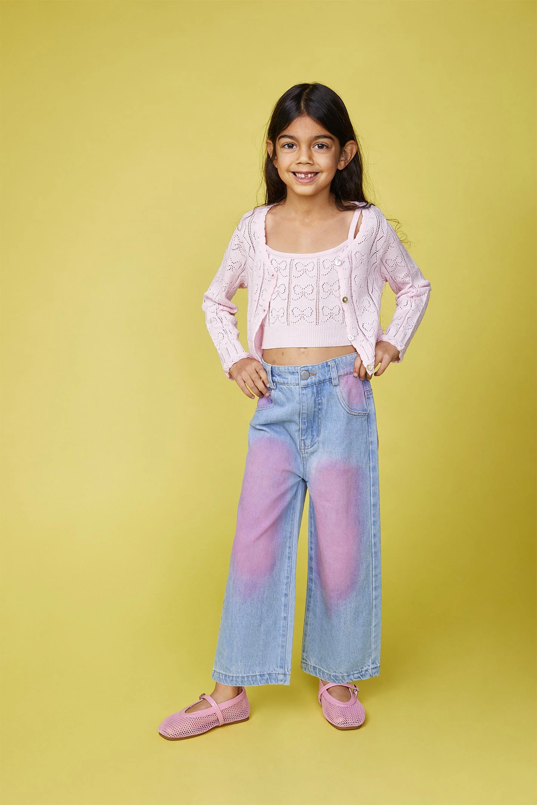 A young girl with dark hair, smiling, standing against a yellow background. She is wearing a pink crocheted cardigan, a white crop top, baggy jeans with pink dye accents, and pink shoes.
