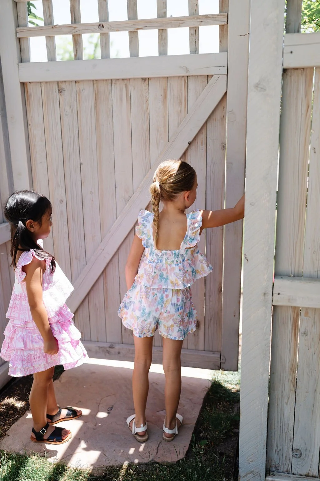 Two young girls standing near a wooden fence gate outdoors. One girl with blonde hair in a braid wearing a floral ruffled dress is opening the gate, while the other girl with long dark hair in a pink dress observes.