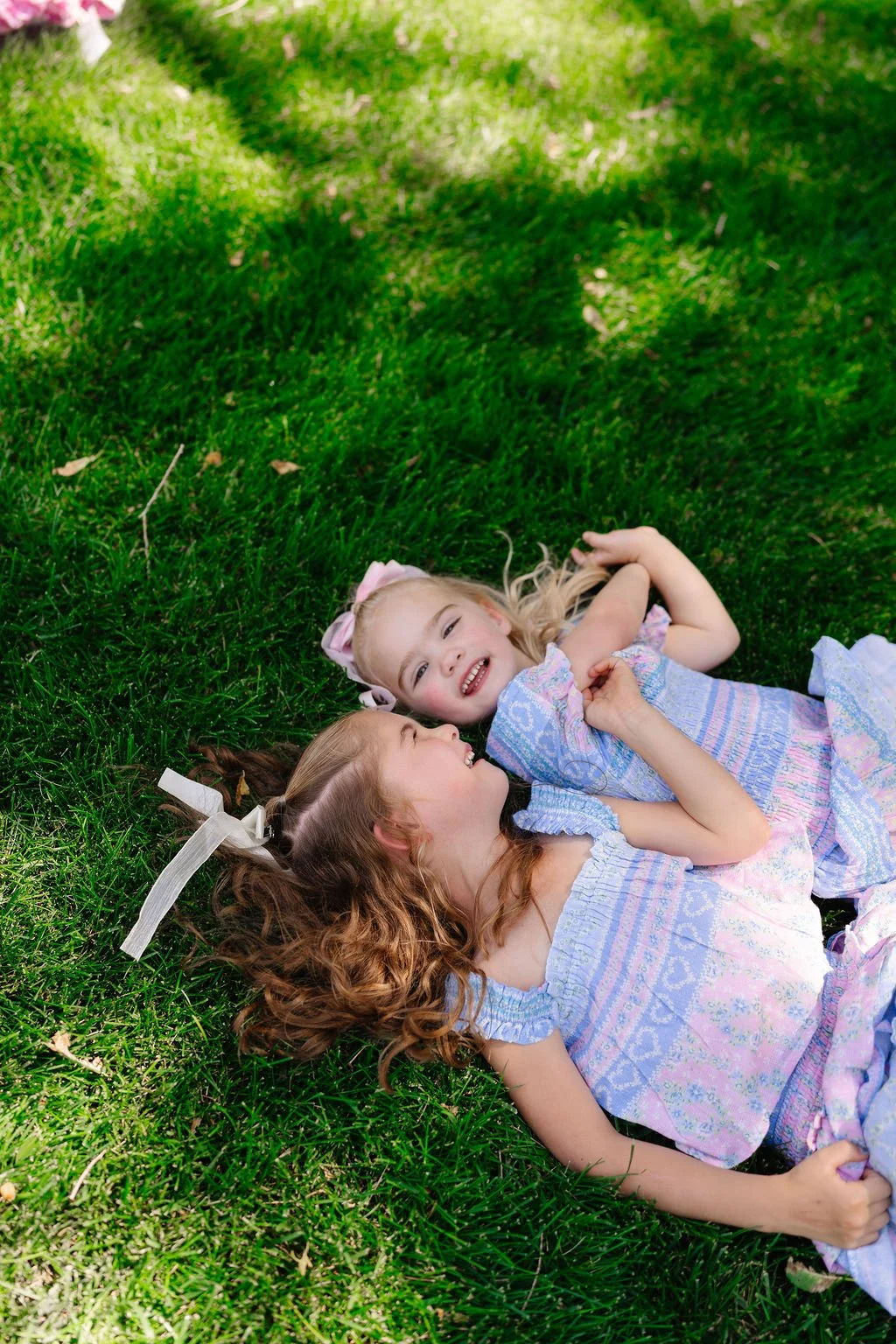 Two smiling young girls lying on green grass, holding hands, and looking at each other.