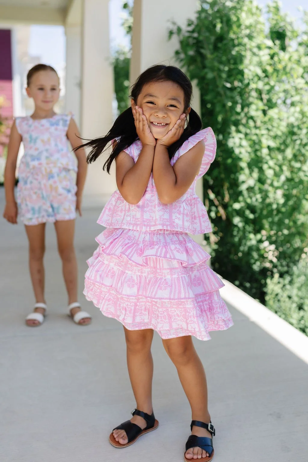Smiling young girl in pink dress with layered ruffles standing outdoors with hands on cheeks, with another girl in the background wearing a colorful dress.
