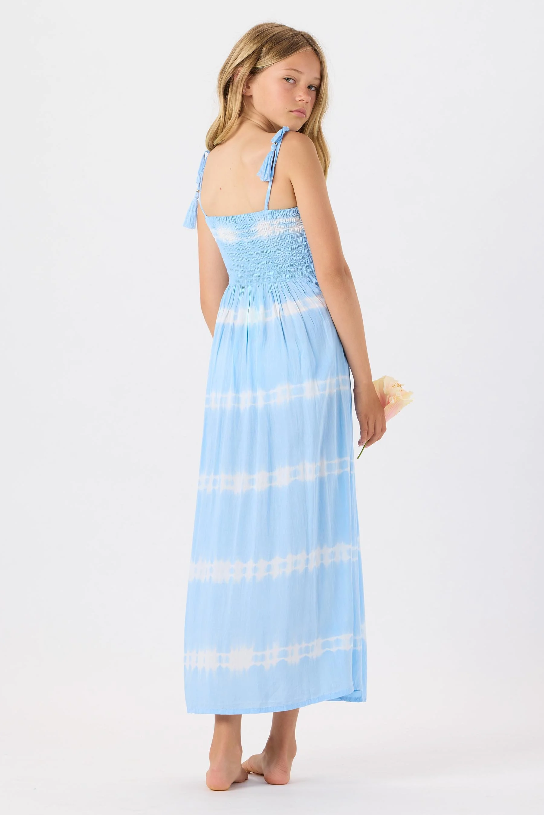 Young girl with long blonde hair wearing a blue tie-dye sleeveless maxi dress with shoulder ties, standing barefoot against a white background, holding a pink flower in her right hand.