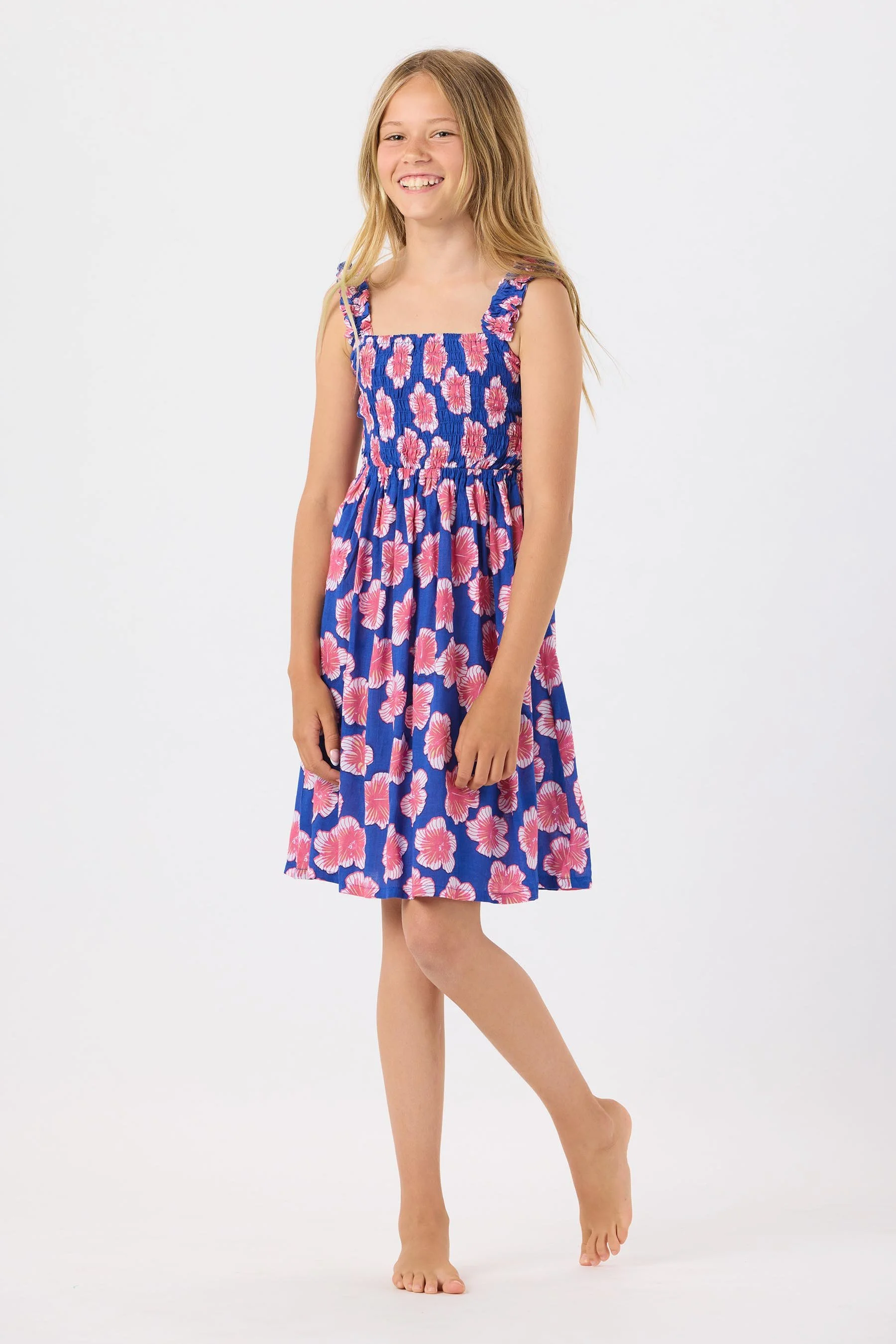 A young girl with long light brown hair smiling, wearing a blue dress with pink floral patterns and ruffled straps, standing barefoot against a white background.