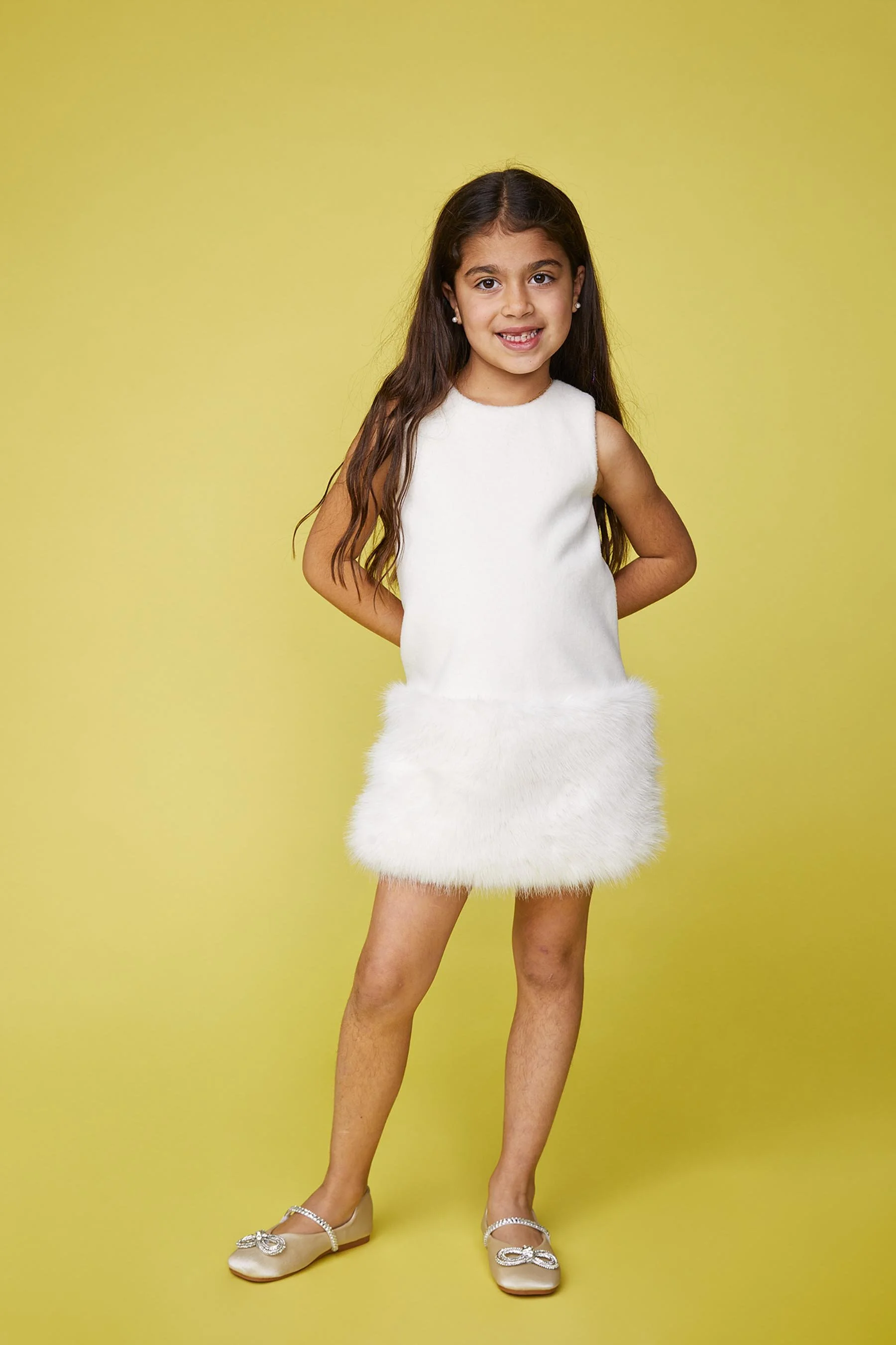 Young girl with long brown hair, wearing a white dress with furry hem, standing against a yellow background, smiling, with her hands behind her back and wearing beaded shoes.