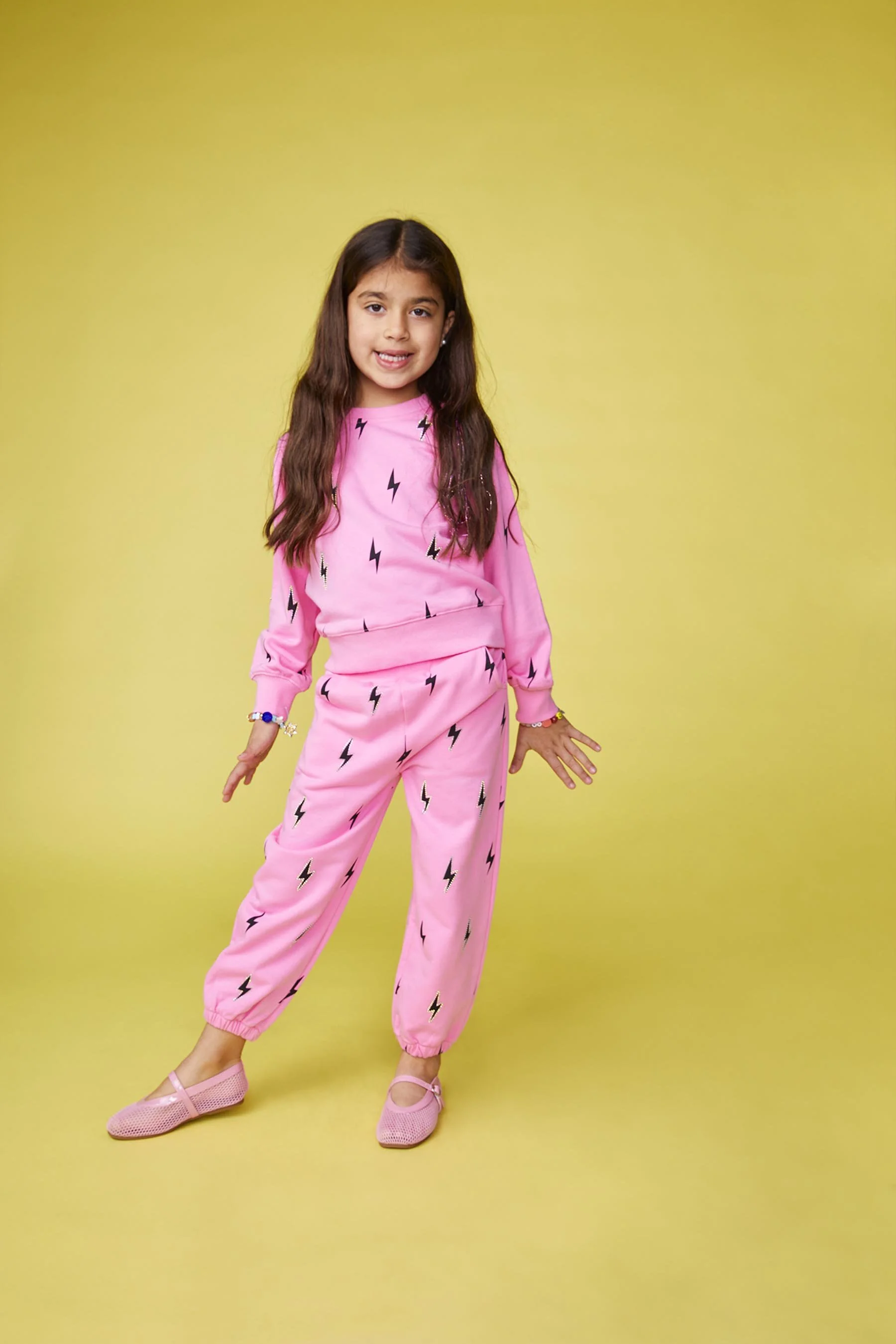 Young girl standing on a yellow background wearing pink pajamas with black lightning bolt patterns and pink shoes.