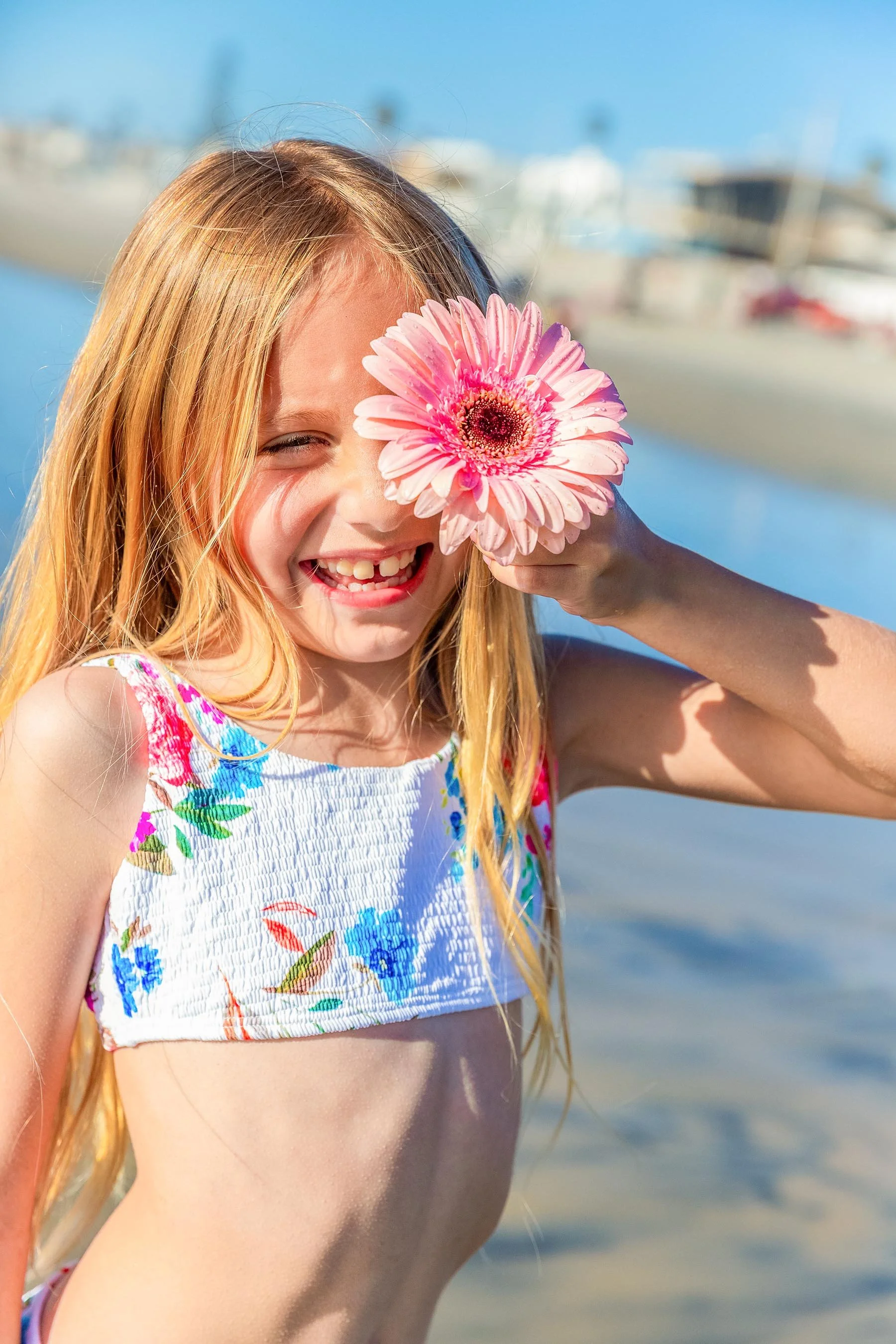 A young girl with long blonde hair smiling and holding a pink flower in front of her face at the beach.