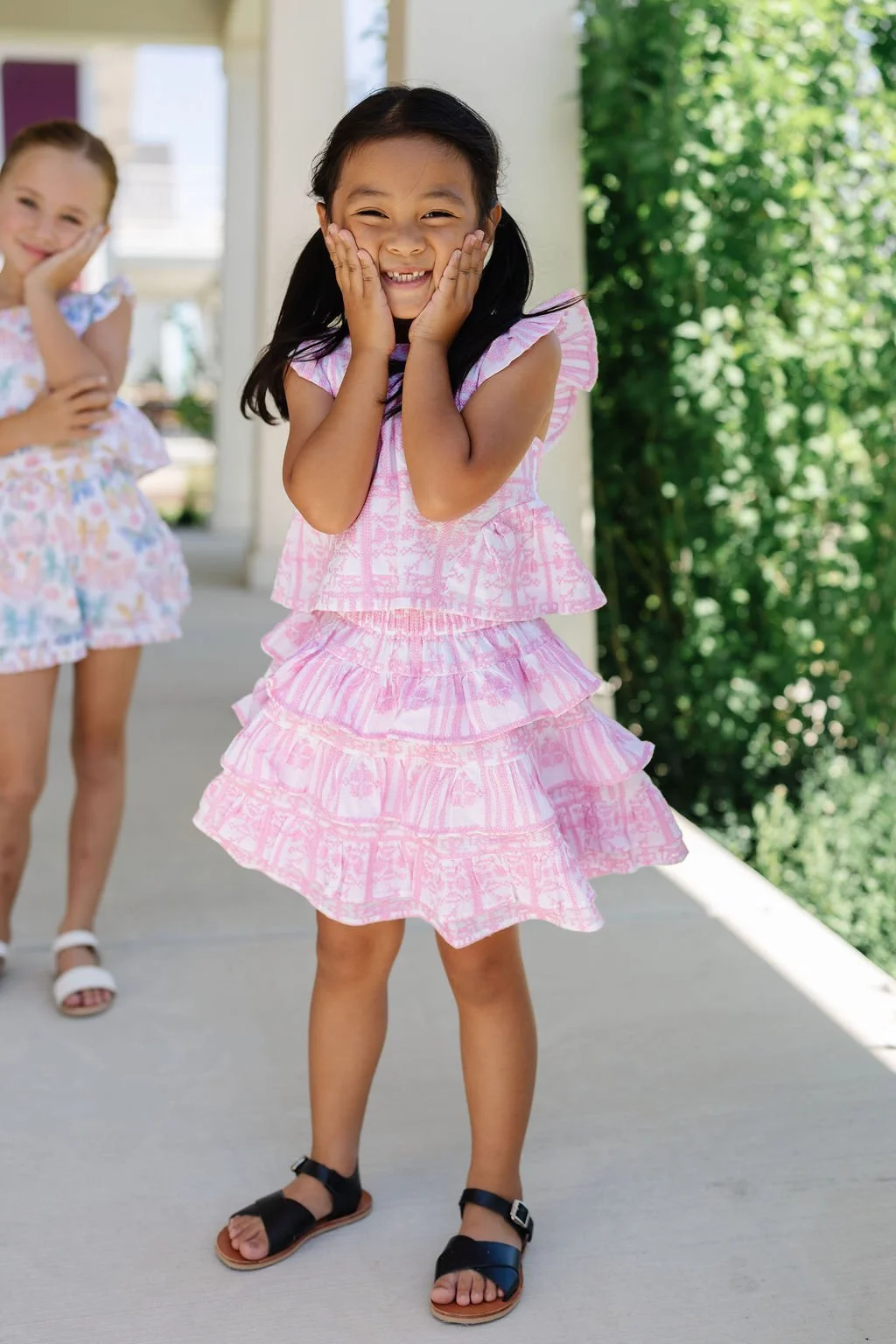 Smiling young girl in a pink dress with ruffles, holding her cheeks, standing outdoors with greenery in the background. Another girl in a colorful dress is in the background.