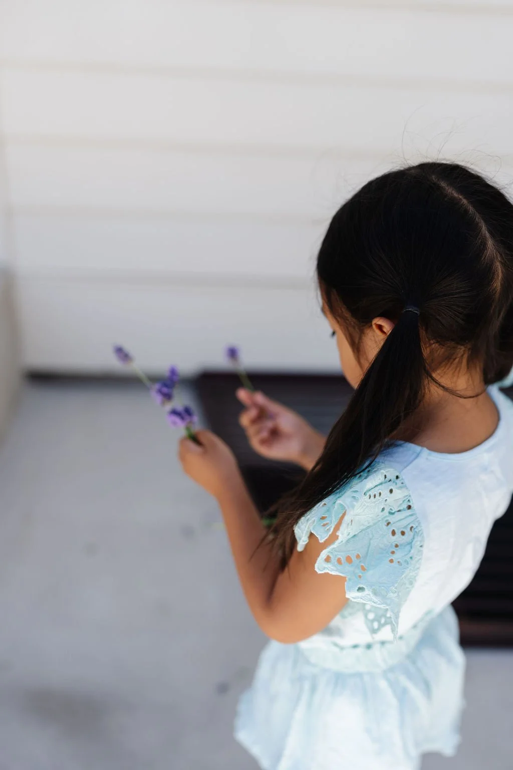 A young girl with dark hair tied in a ponytail, wearing a light blue dress with eyelet details, holding purple flowers, standing in front of a white wall.