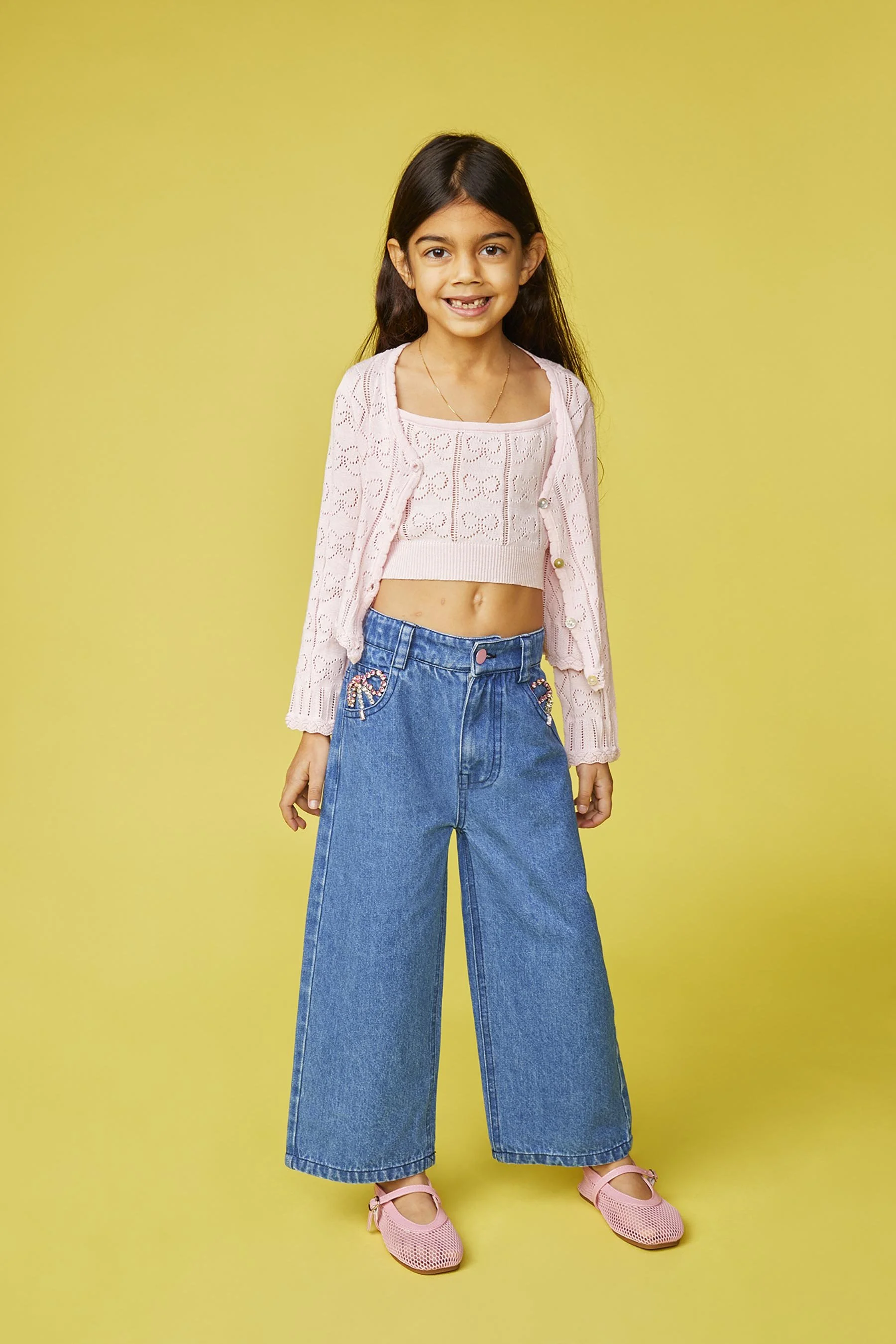 Young girl with long dark hair, wearing a pink lace crop top, pink cardigan, wide-leg jeans with decorated pockets, and pink shoes, standing against a yellow background.