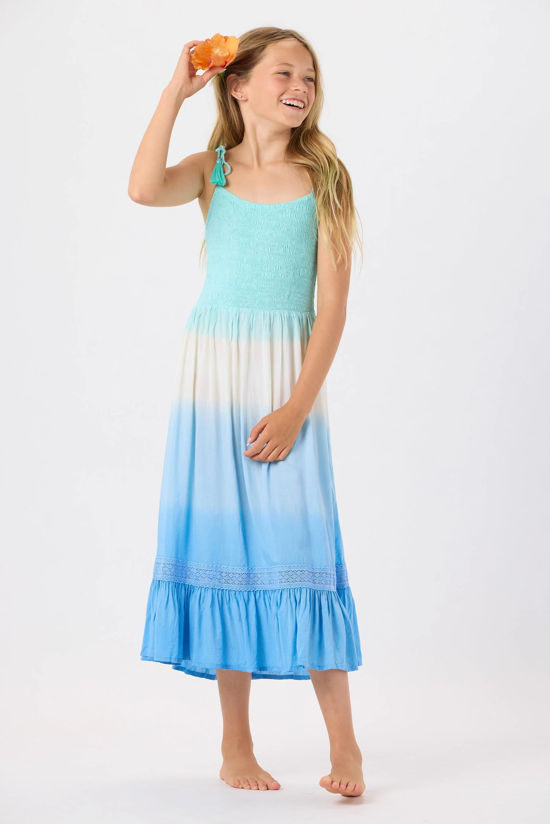 A young girl with long blonde hair, wearing a sleeveless maxi dress with a turquoise top and a gradient blue and white skirt, holding an orange flower to her head and smiling.