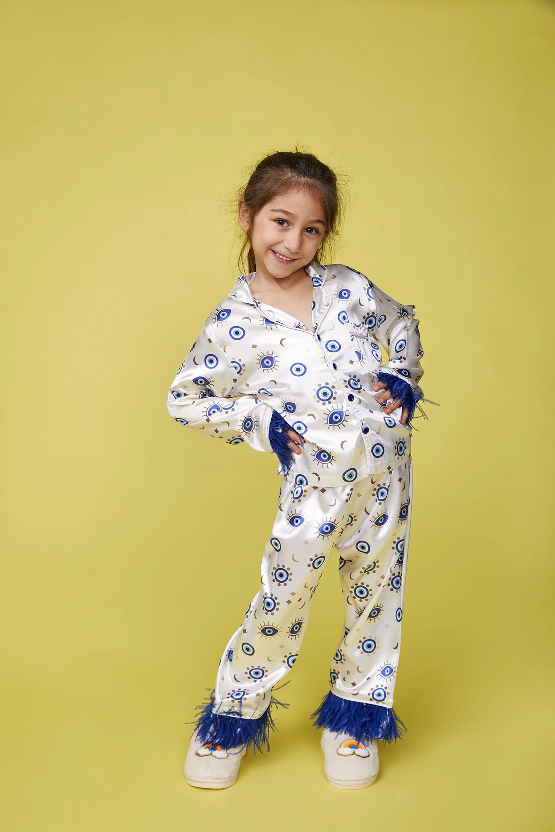 A young girl with brown hair in a ponytail, wearing cream-colored pajamas with blue eye patterns and feathery blue trim on the sleeves and pant cuffs, standing against a yellow background with a smile and hands in her pockets.