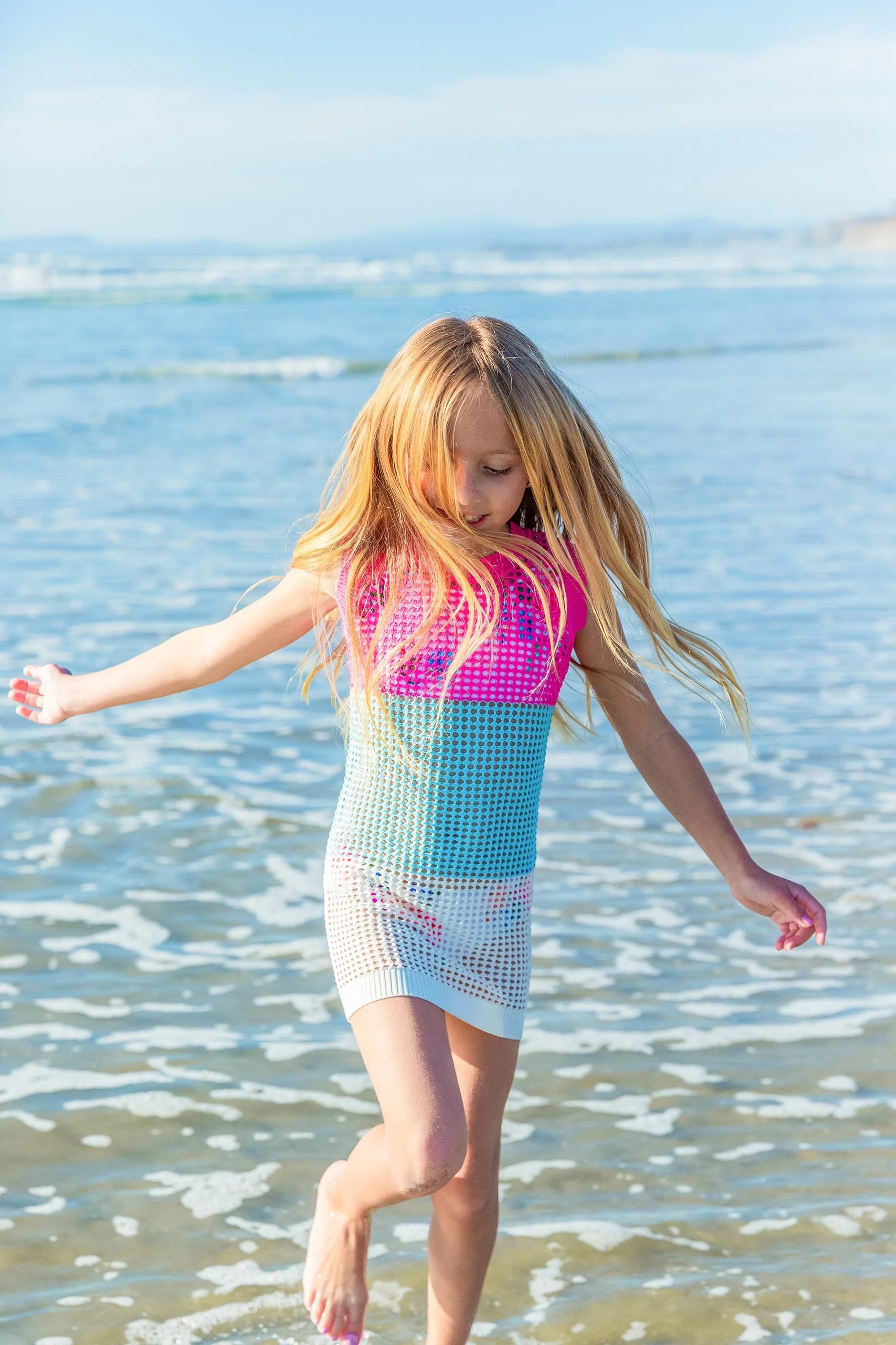A young girl with long blonde hair is playing in the shallow water at the beach, wearing a colorful knitted dress.