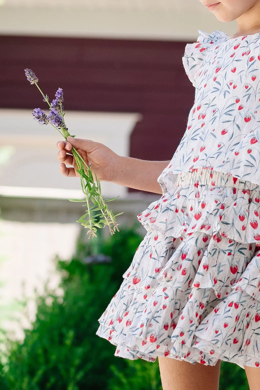 A young girl holding a small bunch of purple lavender flowers in her hand, wearing a white dress with a cherry and leaf pattern and ruffled layers.