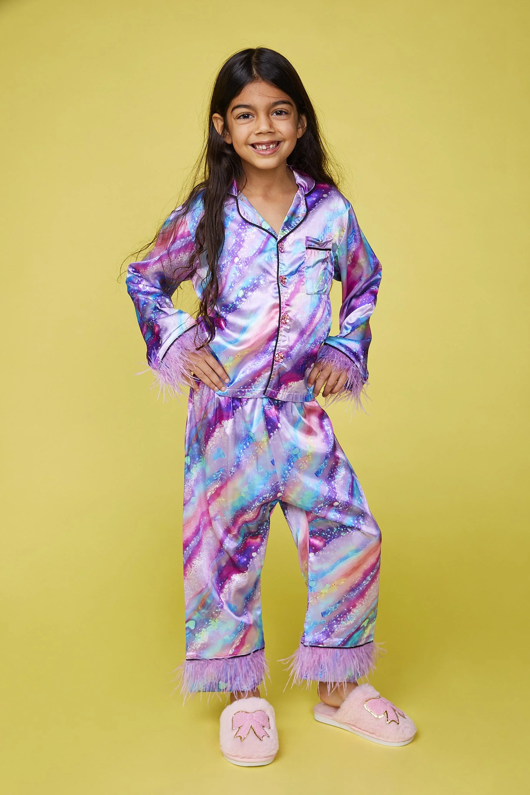 A young girl standing with her hands on her hips, wearing colorful galaxy-patterned pajamas with pink feather trim and pink slippers with a bow design, against a yellow background.