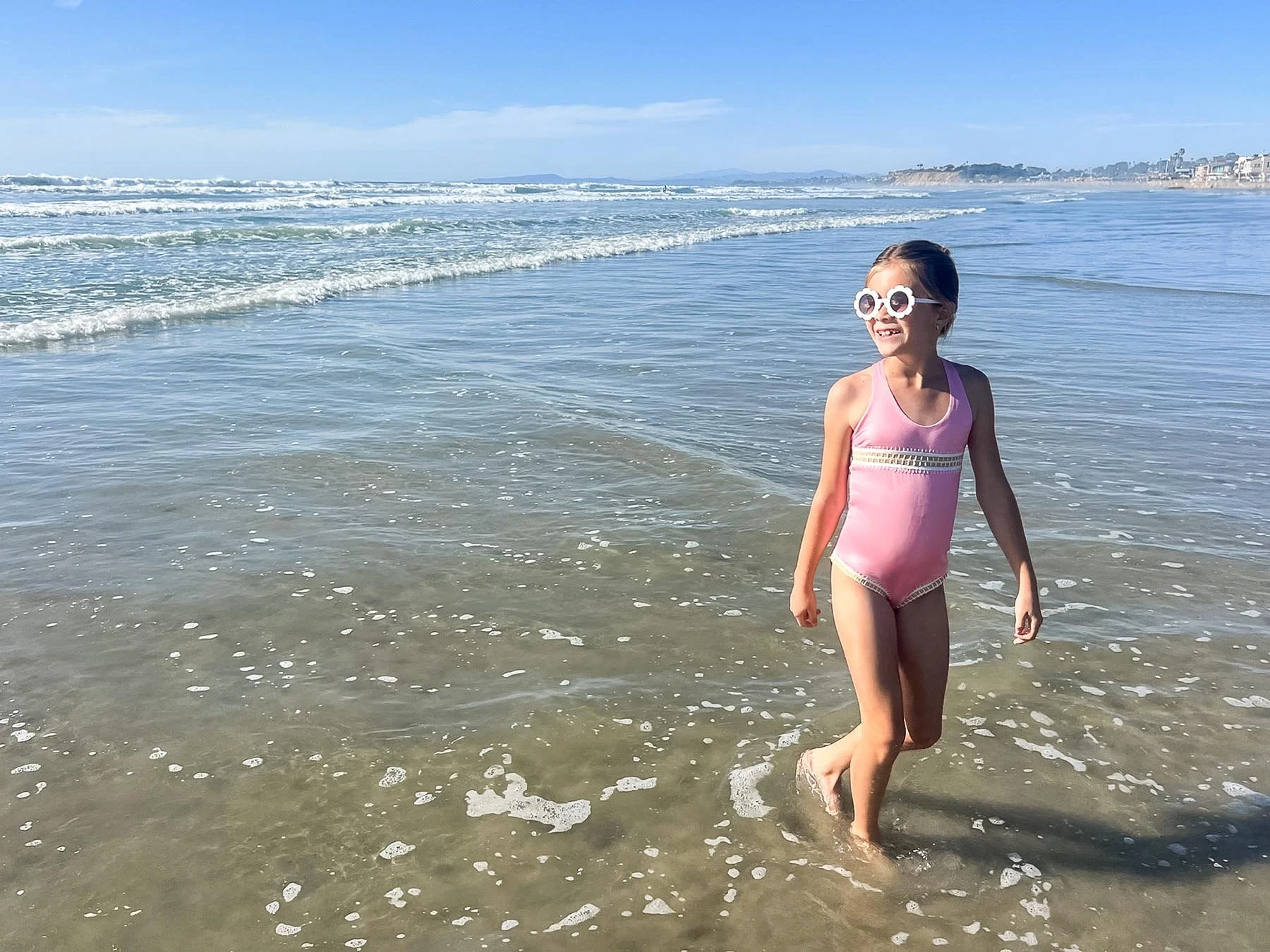 A young girl wearing a pink swimsuit and white sunglasses with flower frames, smiling and walking in shallow ocean water at the beach on a sunny day.