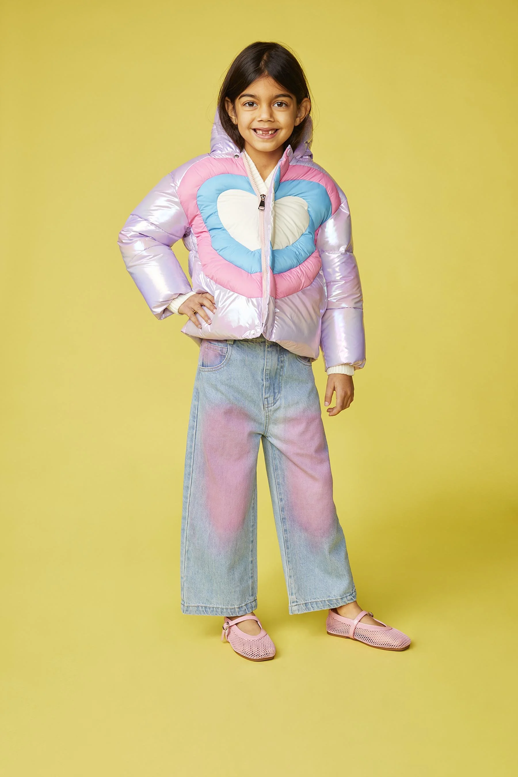 A young girl with dark hair, smiling, wearing a shiny, pink and blue puffer jacket with a heart detail, baggy jeans with pink tie-dye patches, pink shoes, standing against a yellow background.