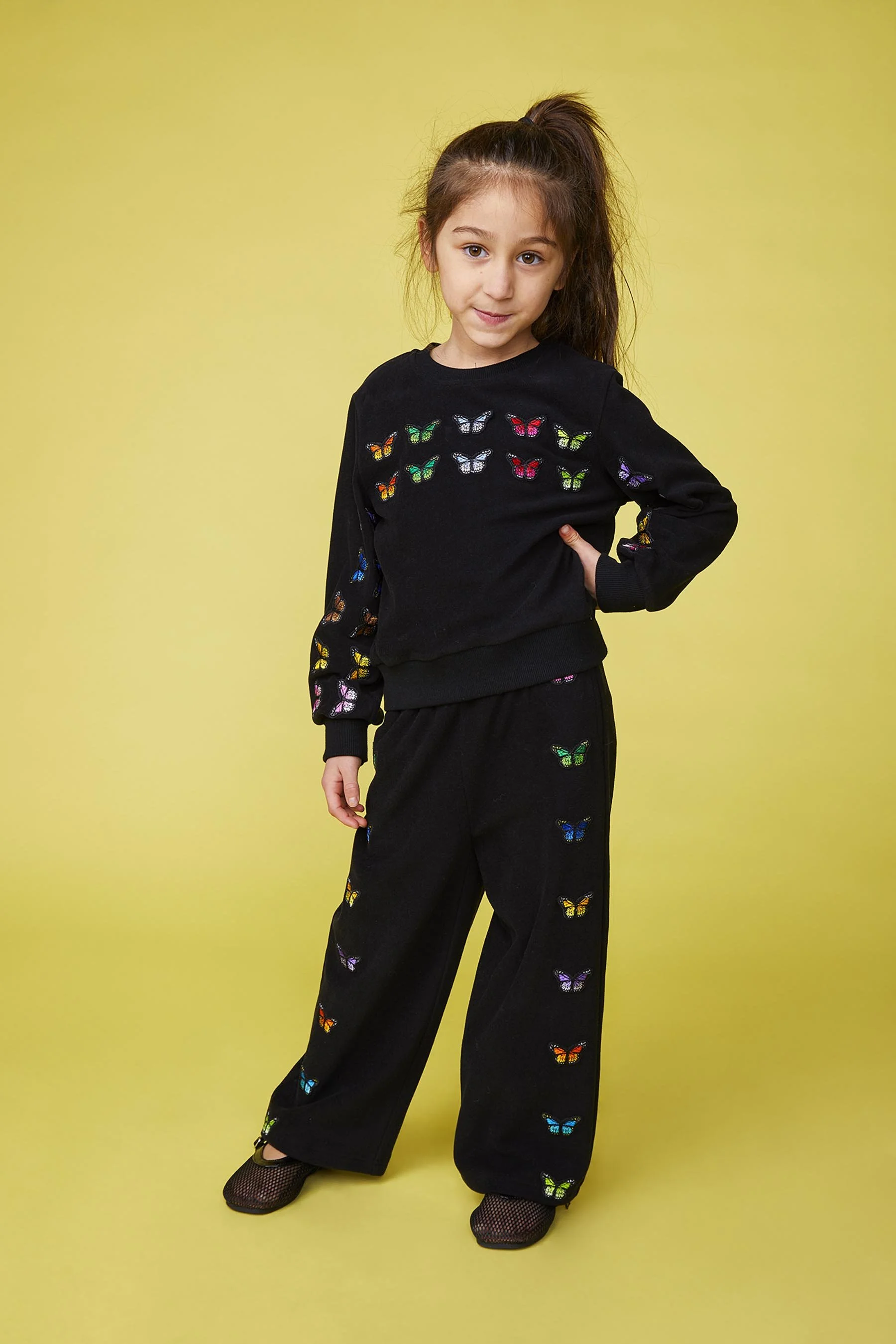 A young girl with brown hair in a high ponytail, posing against a yellow background. She is wearing a black outfit decorated with colorful butterfly patterns.