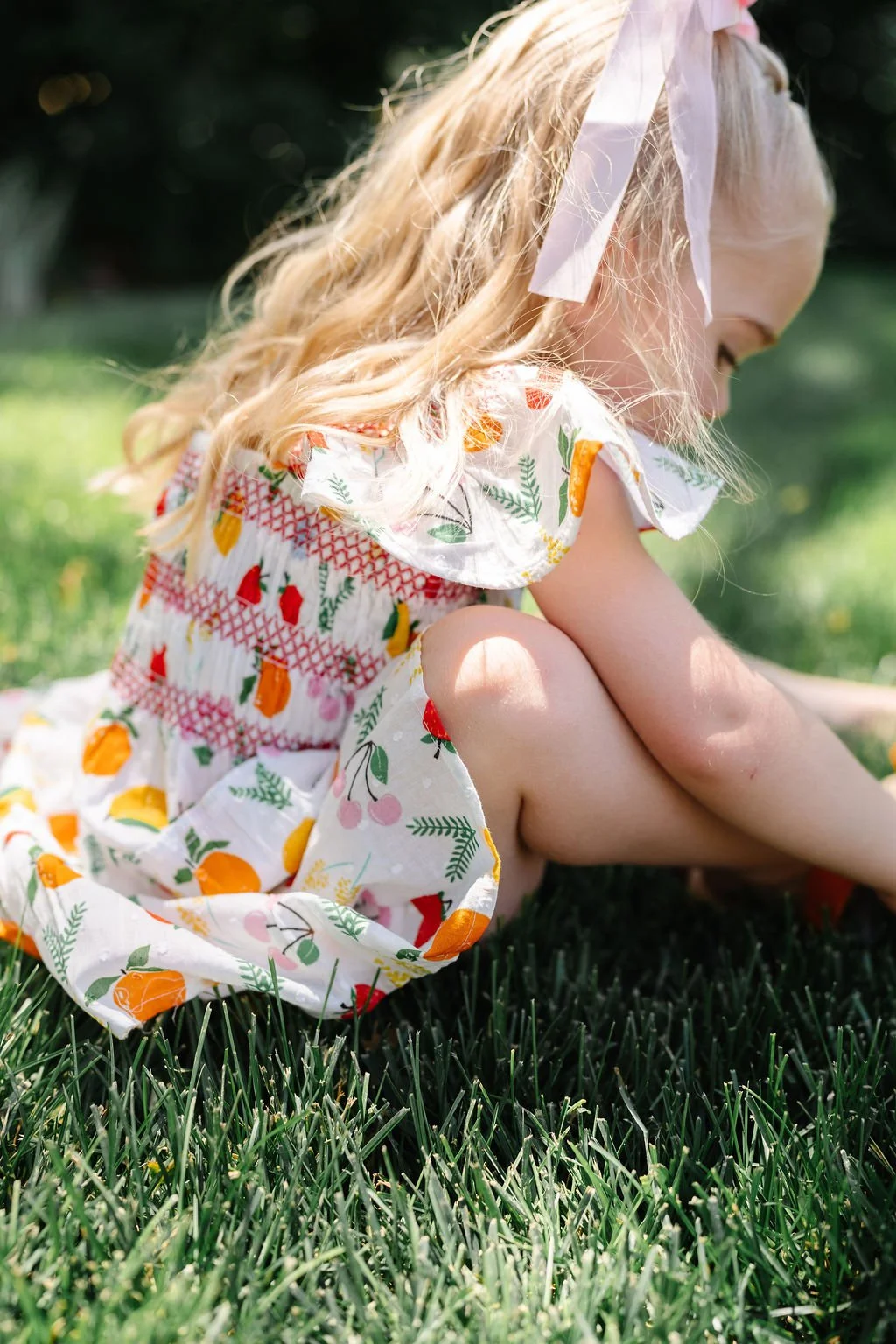 A young girl with blonde curly hair sitting on grass outdoors, wearing a colorful dress with fruit patterns and a white ribbon in her hair.
