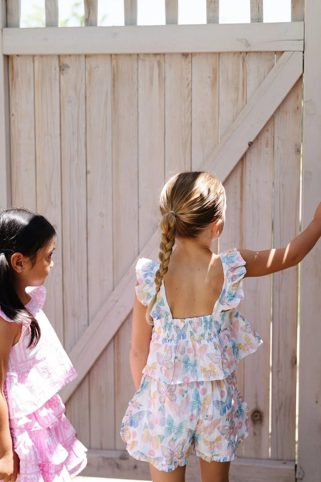 Two young girls wearing pastel-colored summer dresses with butterfly patterns standing in front of a wooden fence, one girl reaching up to open or close the gate.