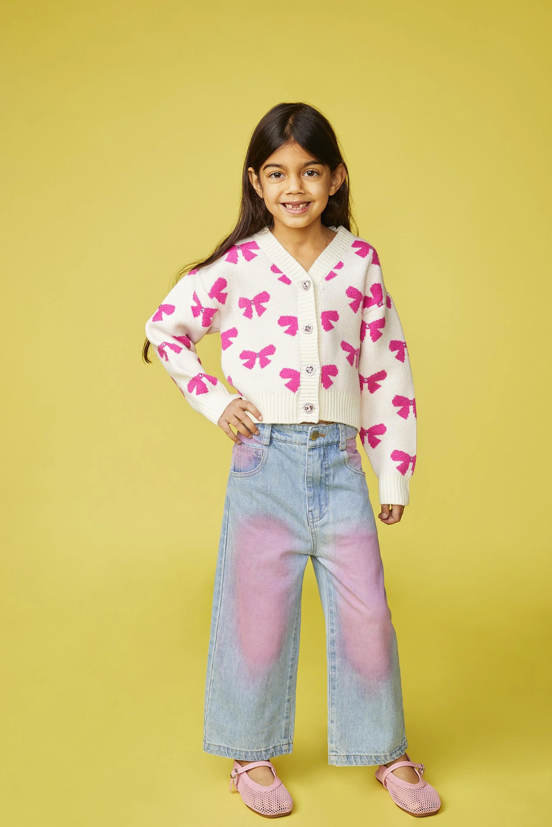 A young girl with long dark hair, smiling, wearing a white sweater with pink bows, light denim pants with pink patches, and pink shoes, standing against a yellow background.