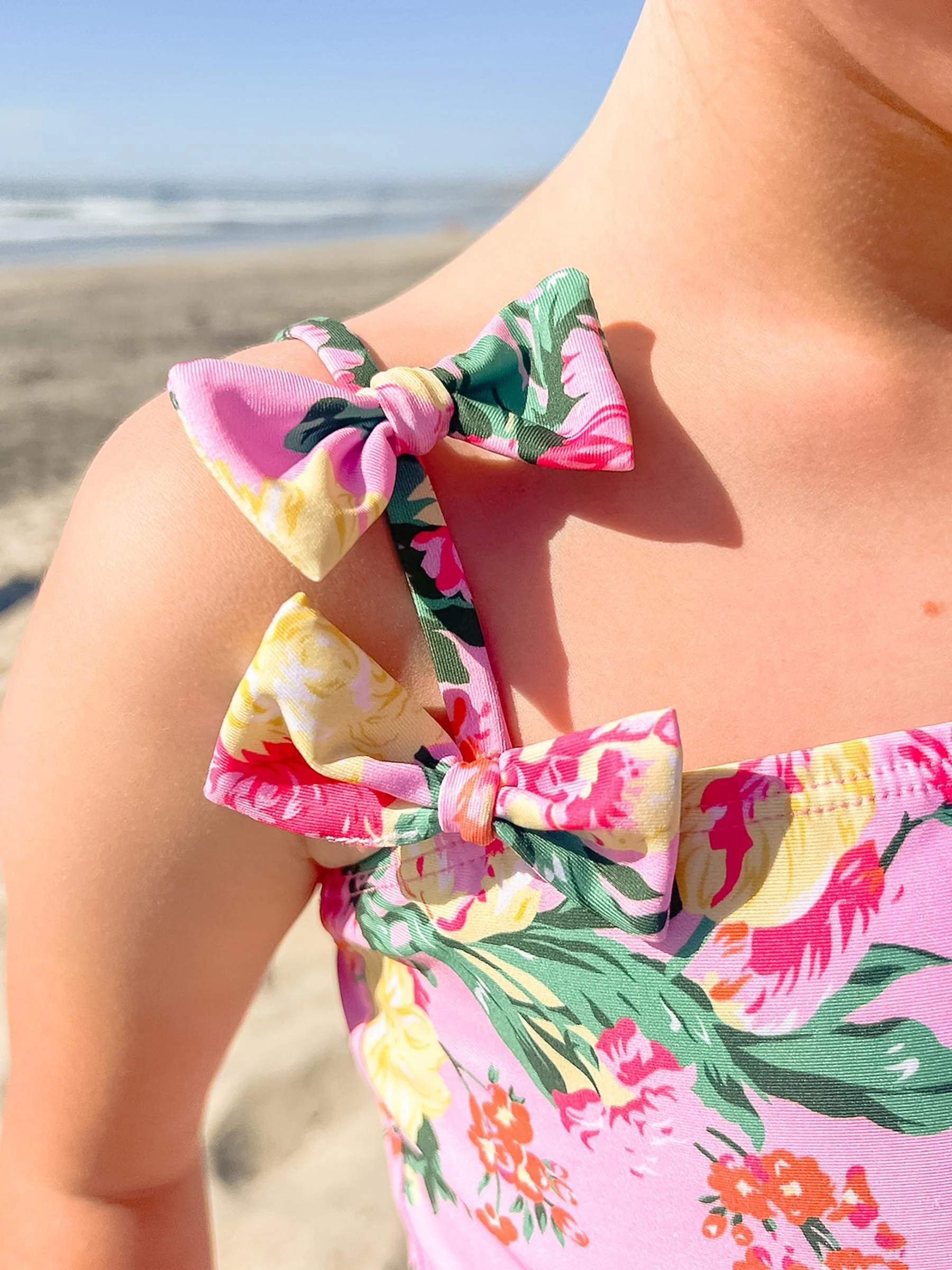 Close-up of a woman's shoulder wearing a pink floral dress with bow details on the straps at the beach.