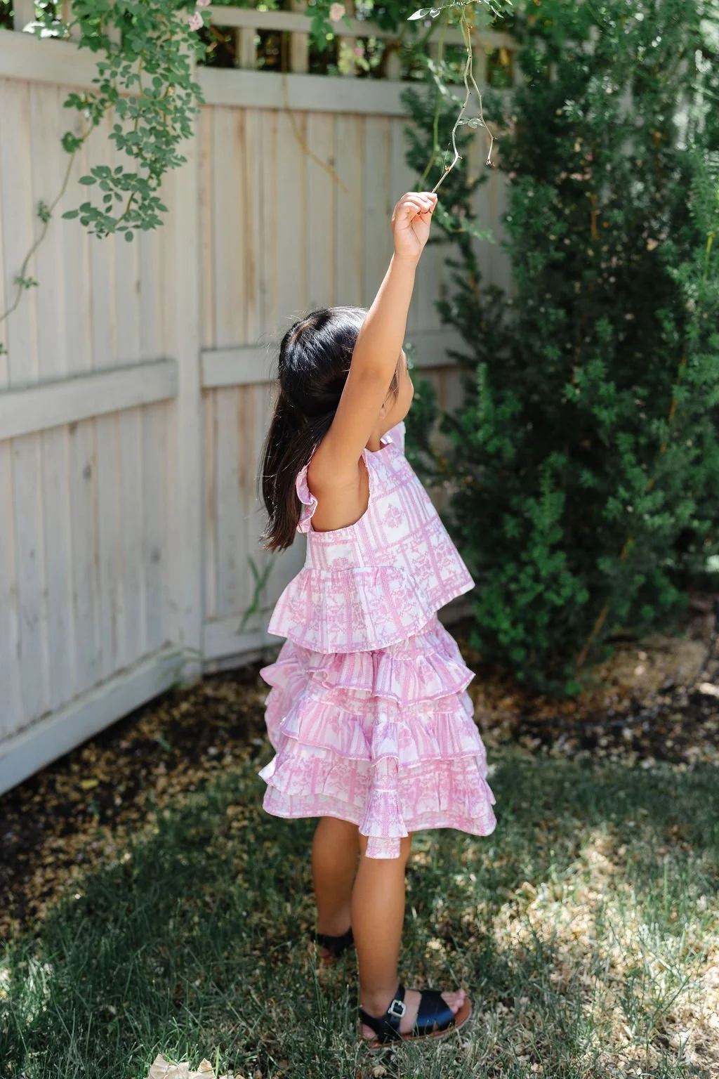 A young girl with long dark hair wearing a pink ruffled dress, standing in a backyard, reaching up to grab a vine hanging from a tree.