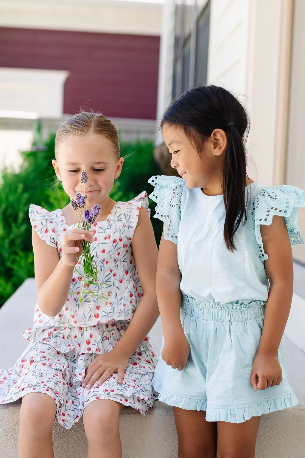 Two young girls, one holding a bouquet of lavender flowers, stand outdoors near a porch with greenery in the background. The girl on the left has her eyes closed as she smells the flowers, and the girl on the right is smiling at her.