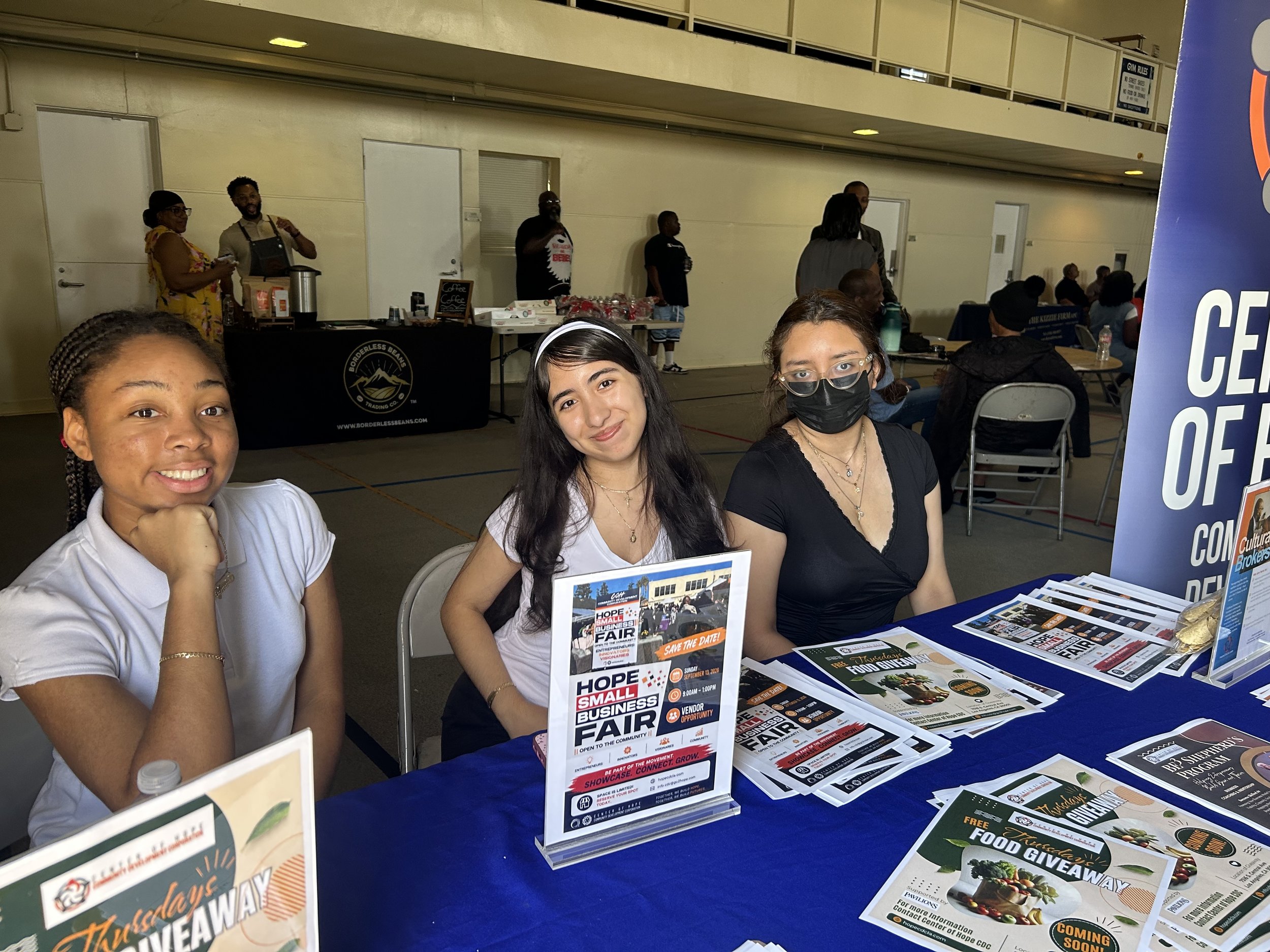 Three young women sitting at a table with flyers and brochures at a small business fair, with a large banner and other people in the background.