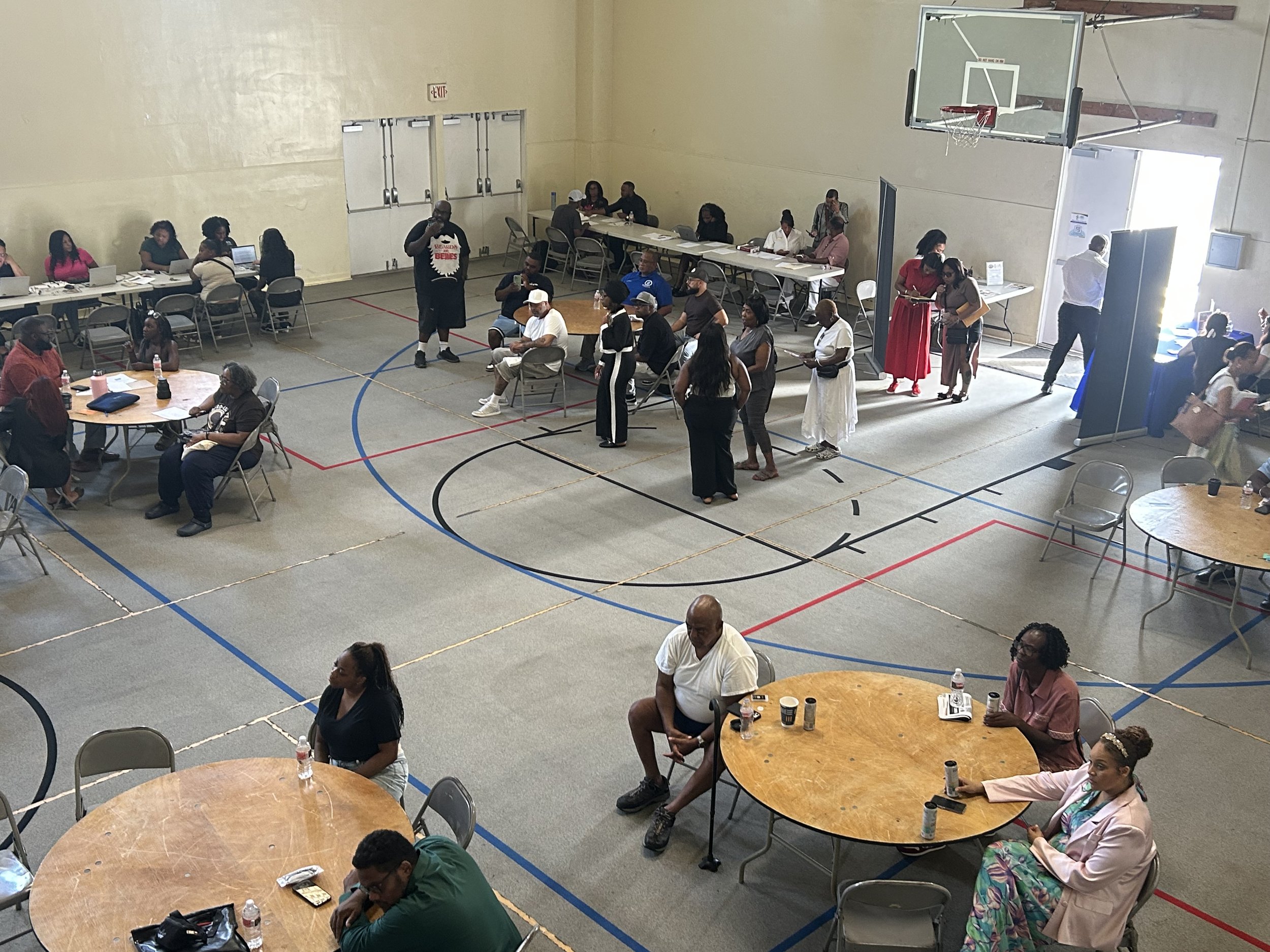 People gathered in a gymnasium at tables, some sitting and talking, others standing or walking around, engaging in a community event or meeting.