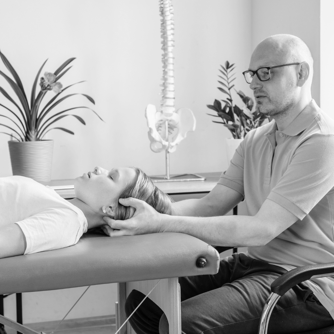 A chiropractor examines a woman's neck while she lies on an examination table, with a skeleton model and potted plants in the background.