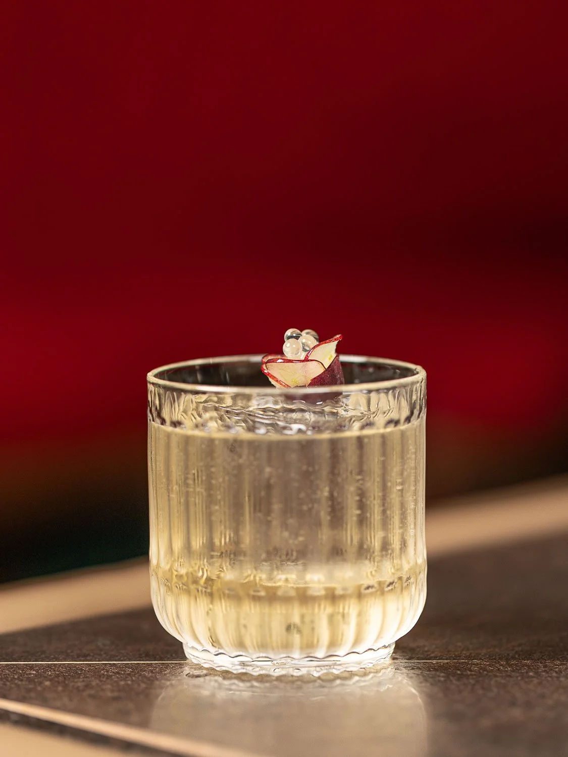 Ein Glas mit einem klaren Getränk, garniert mit einer dünnen Scheibe roter Zwiebel und weißen Perlen, auf einer dunklen Oberfläche, mit rotem Hintergrund.
