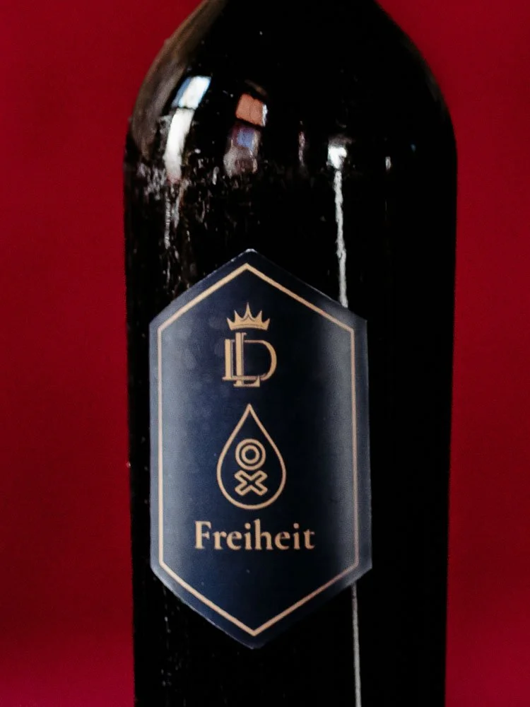 Schwarze Flasche vor rotem Hintergrund, auf dem schwarzen Label ist das Liquid Diary Logo, ein Oxymel-Zeichen und die Aufschrift "Freiheit" zu sehen.