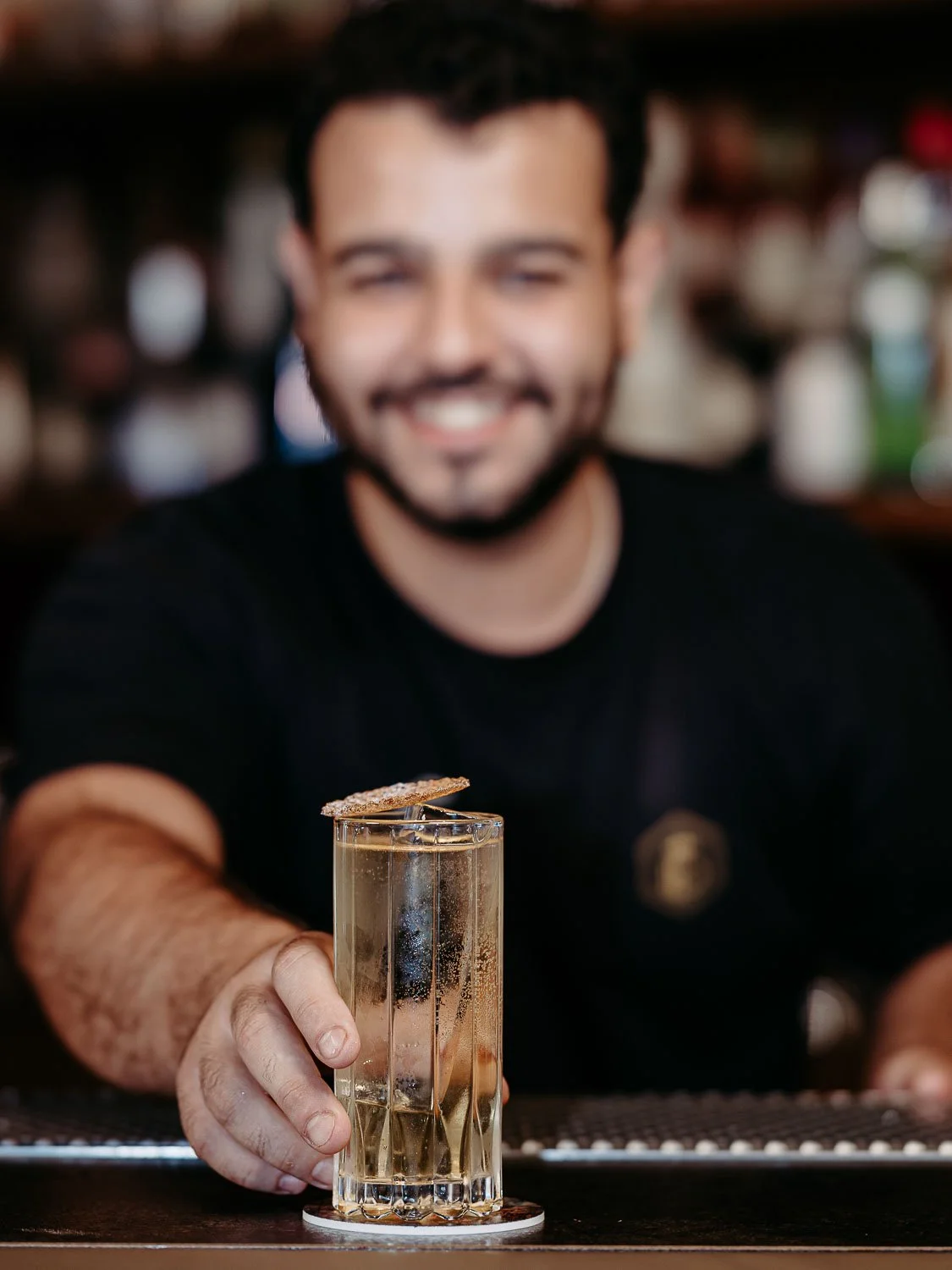 Ein Mann hält ein Glas mit einem Getränk, das mit einem Löffel und einem Keks dekoriert ist, während er lächelnd in einer Bar sitzt.