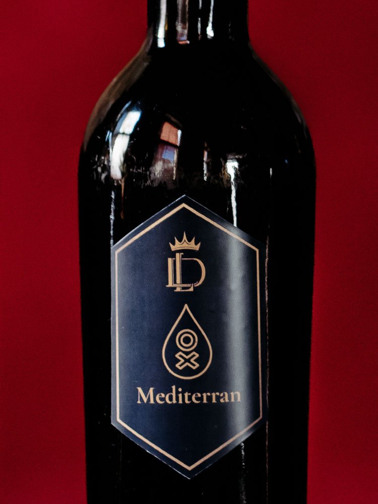 Schwarze Flasche vor rotem Hintergrund, auf dem schwarzen Label ist das Liquid Diary Logo, ein Oxymel-Zeichen und die Aufschrift "Mediterran" zu sehen.