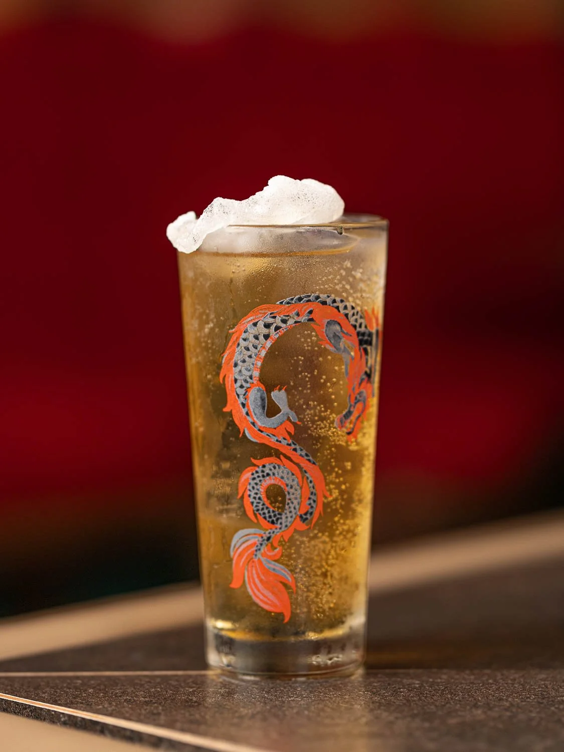 Ein hohes Cocktailglas mit einem aufgedruckten Drachen, darin ein sprudeliger gelber Cocktail und ein langer Eiswürfel garniert mit einem Reischip