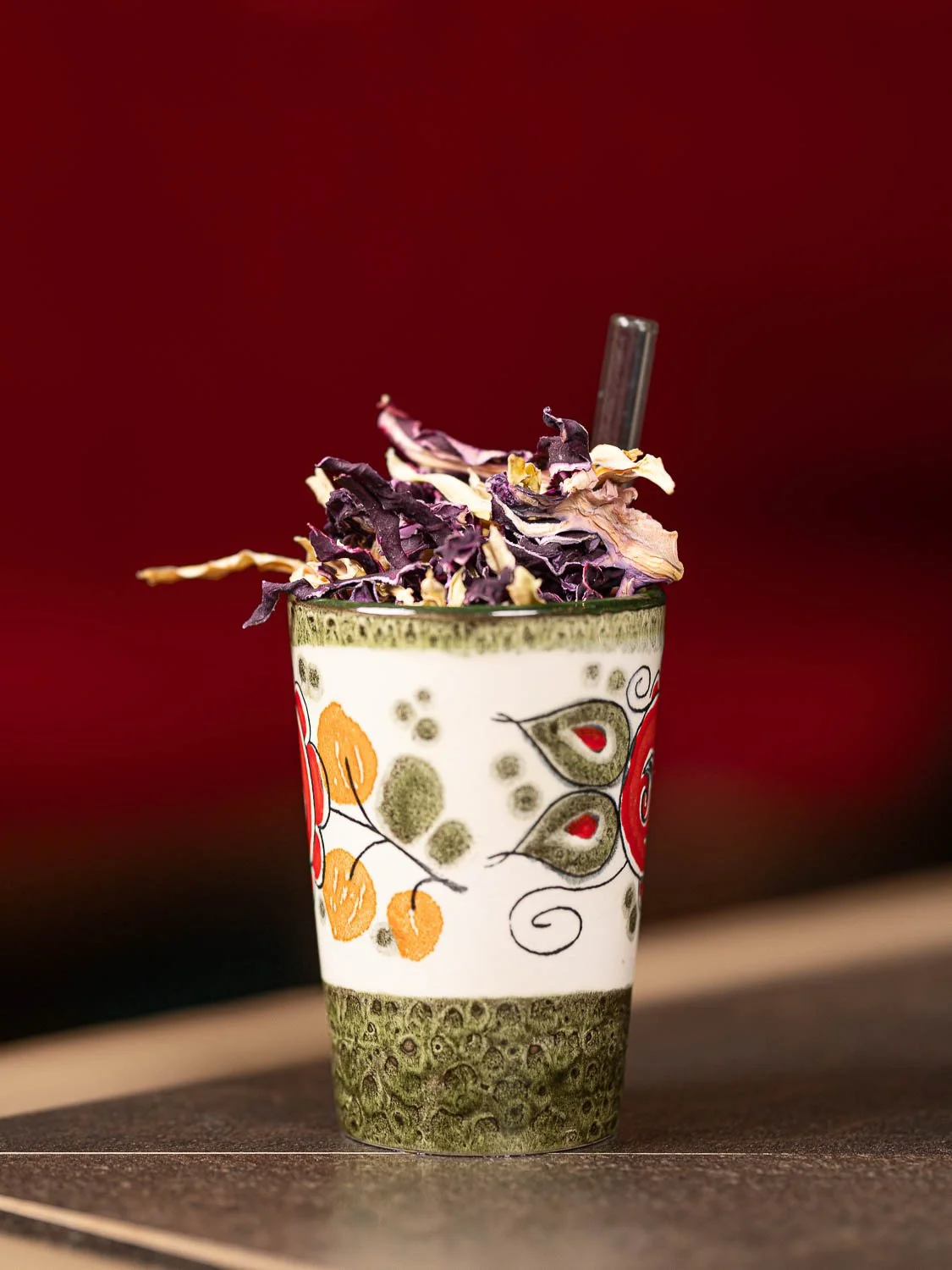 Ein Cocktail in einem bunt bemalten Keramikbecher mit dehydriertem Kraut als Topping in Glas-Strohalm
