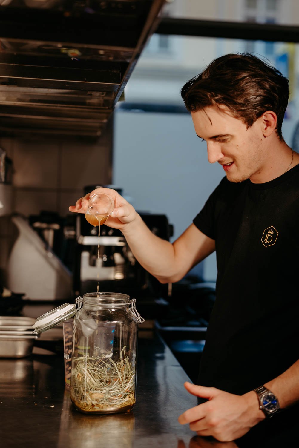 Martin Šiprak, “Head of Oxymel” im Liquid Diary in Innsbruck, setzt ein Oxymel in einem großen Einmachglas an: Er gießt Honig in ein gläsernes Behältnis, das bereits mit Botanicals gefüllt ist.