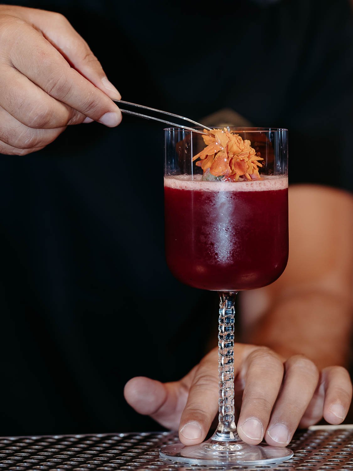Ein dunkelroter Cocktail in einem Glas mit Stil, eine Hand legt eine organgene Blüte oben drauf.