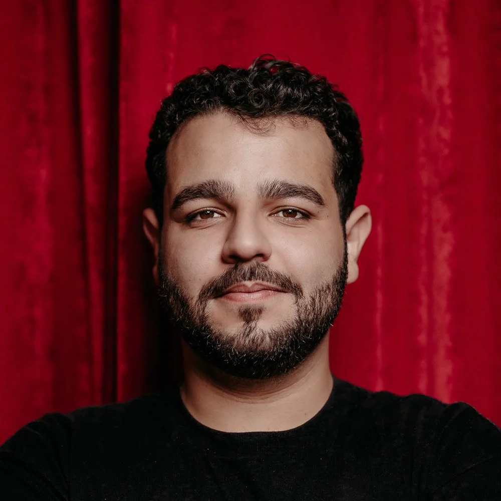 Humberto, ein charismatischer Barkeeper mit Dreitagesbart mit souveränem Blick vor einem rotem Vorhang