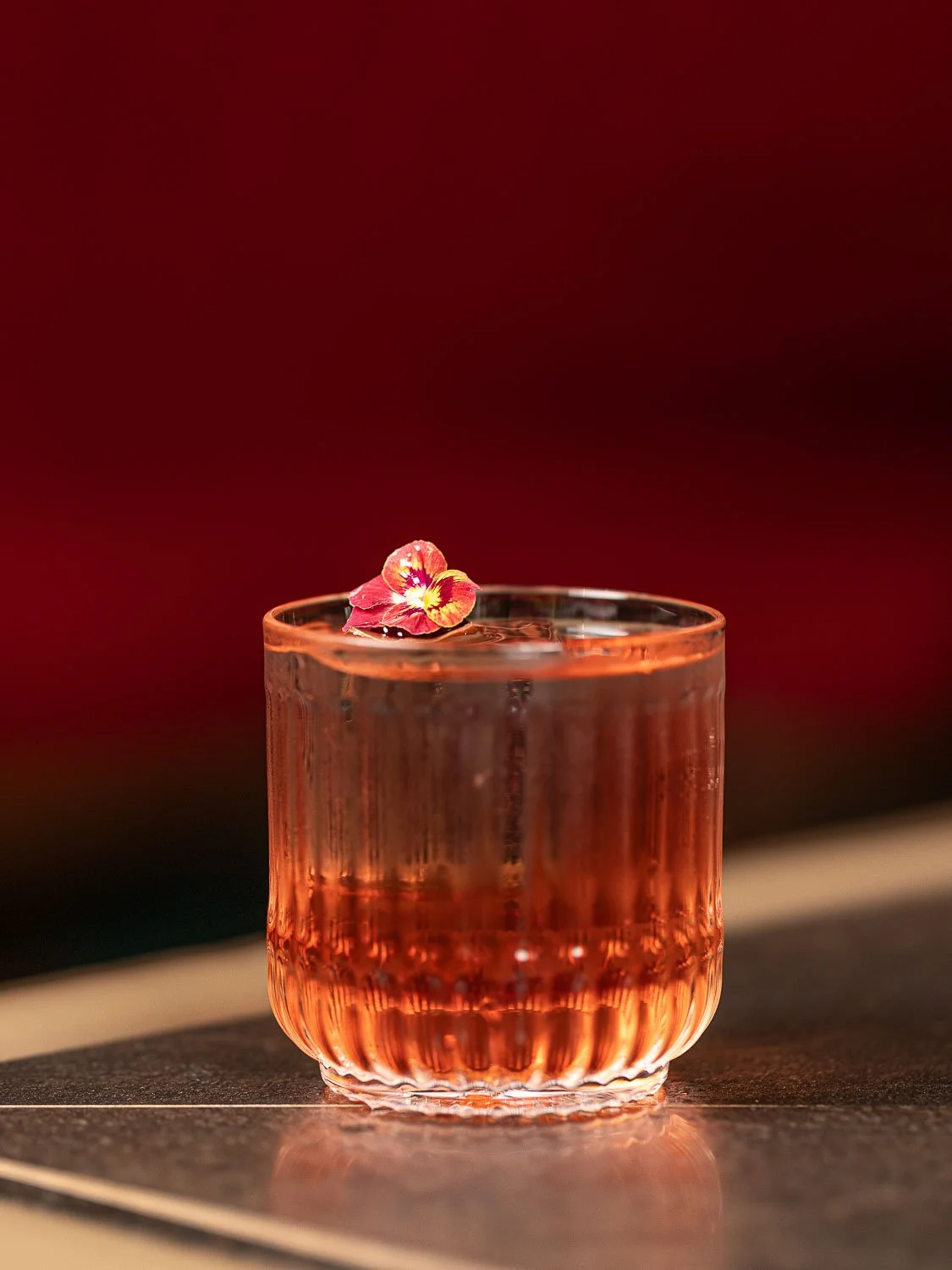 Roter Cocktail in einem hochwertigen Glas mit einer Blüte, die auf dem Eiswürfel liegt.