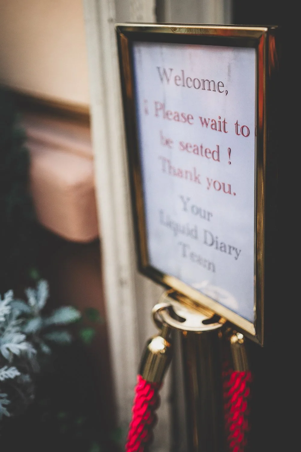 Ein schwarzer Rahmen auf einem goldfarbenen Stativ mit einem weißen Schild, auf dem steht: "Welcome, Please wait to be seated! Thank you. Your Liquid Diary Team".