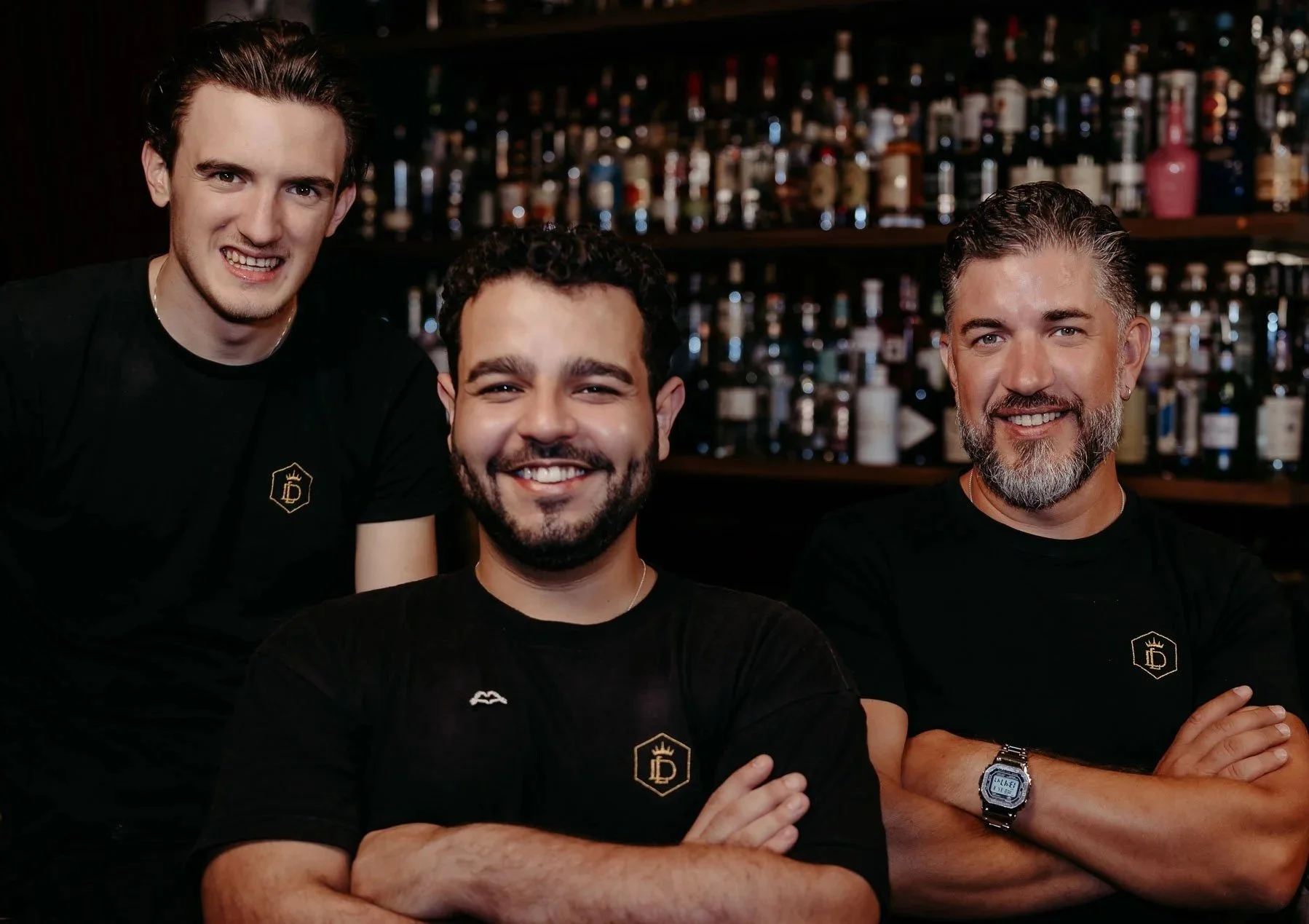 Drei Barkeeper mit schwarzen T-Shirts stehen vor einer Back-Bar mit vielen Flaschen und lächeln in die Kamera.